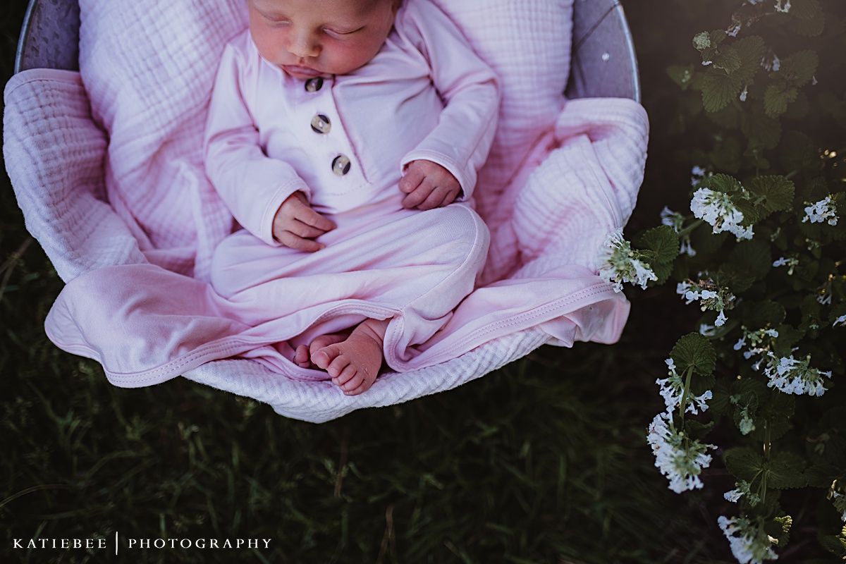 Newborn in pink sleeper rests peacefully in white cushioned basket outdoors.