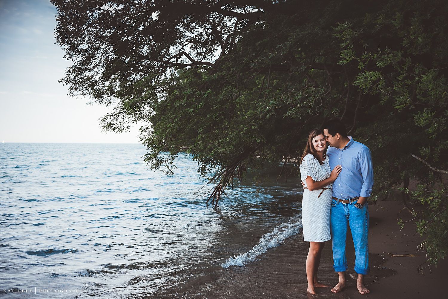 A romantic couple embraces on a beach with waves and trees in the background.