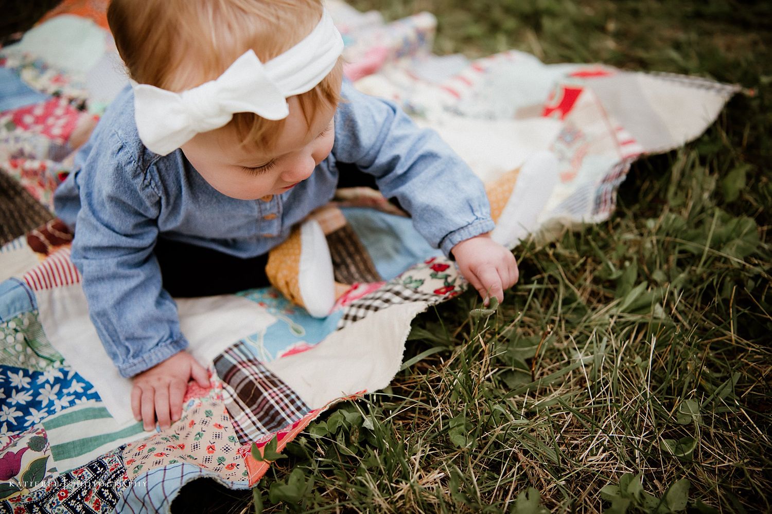 Baby in blue outfit and white bow examining a colorful patchwork quilt on grass.
