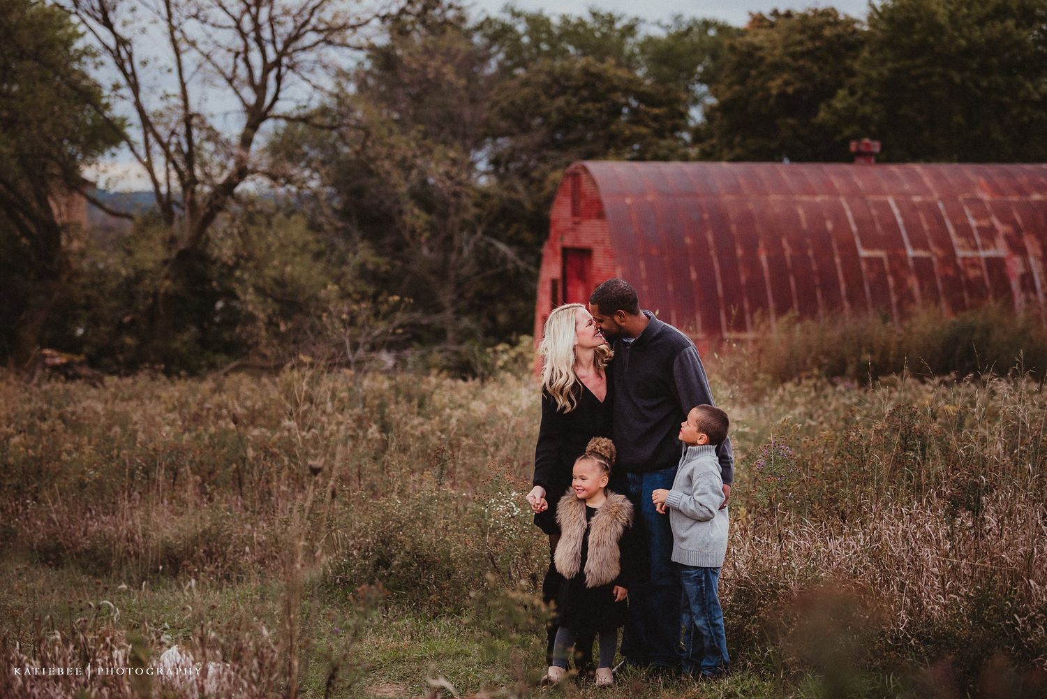 A family portrait taken in front of a rustic red barn surrounded by autumn foliage.