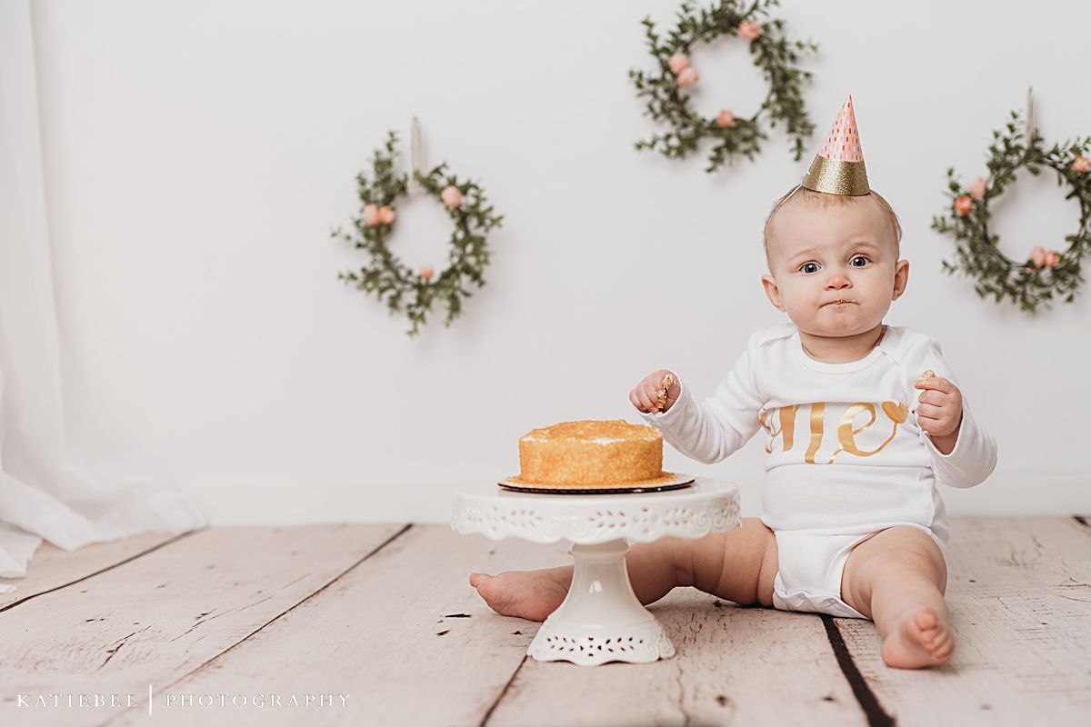 Baby in white outfit with party hat sits near birthday cake with floral wreaths on wall.