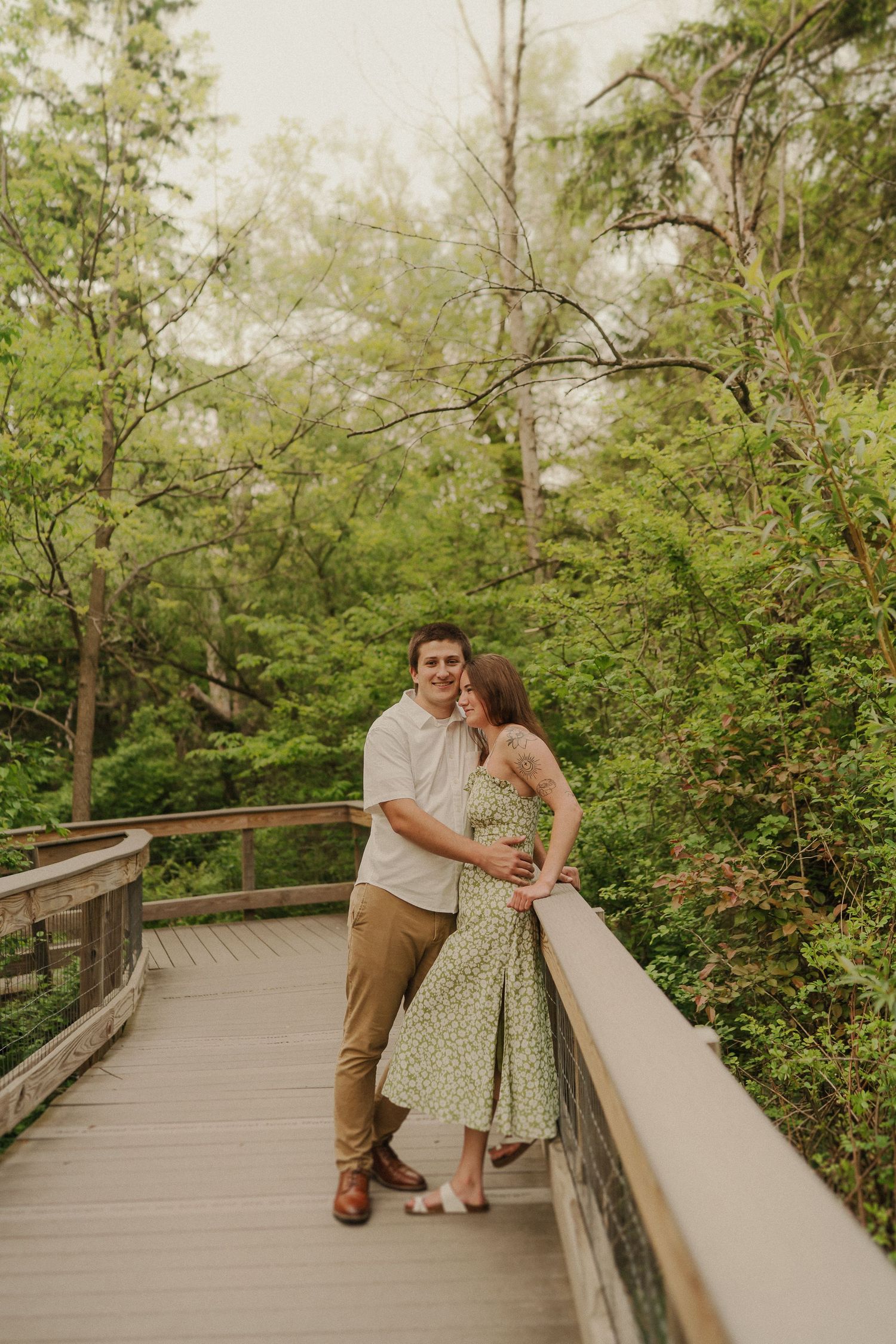 A couple shares an intimate moment on a wooden boardwalk surrounded by lush green foliage.