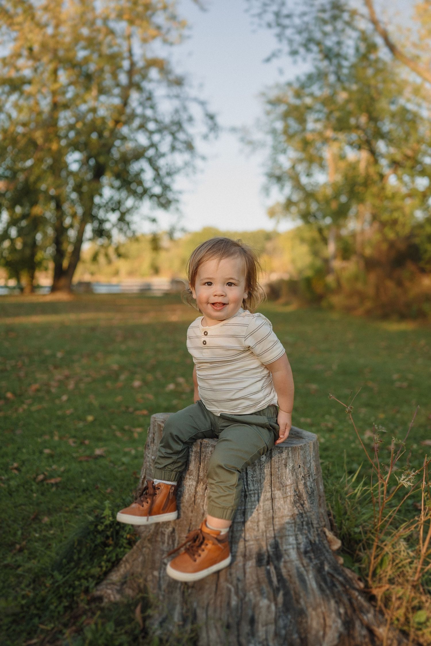 Young child sits happily on tree stump wearing brown boots in autumn setting.