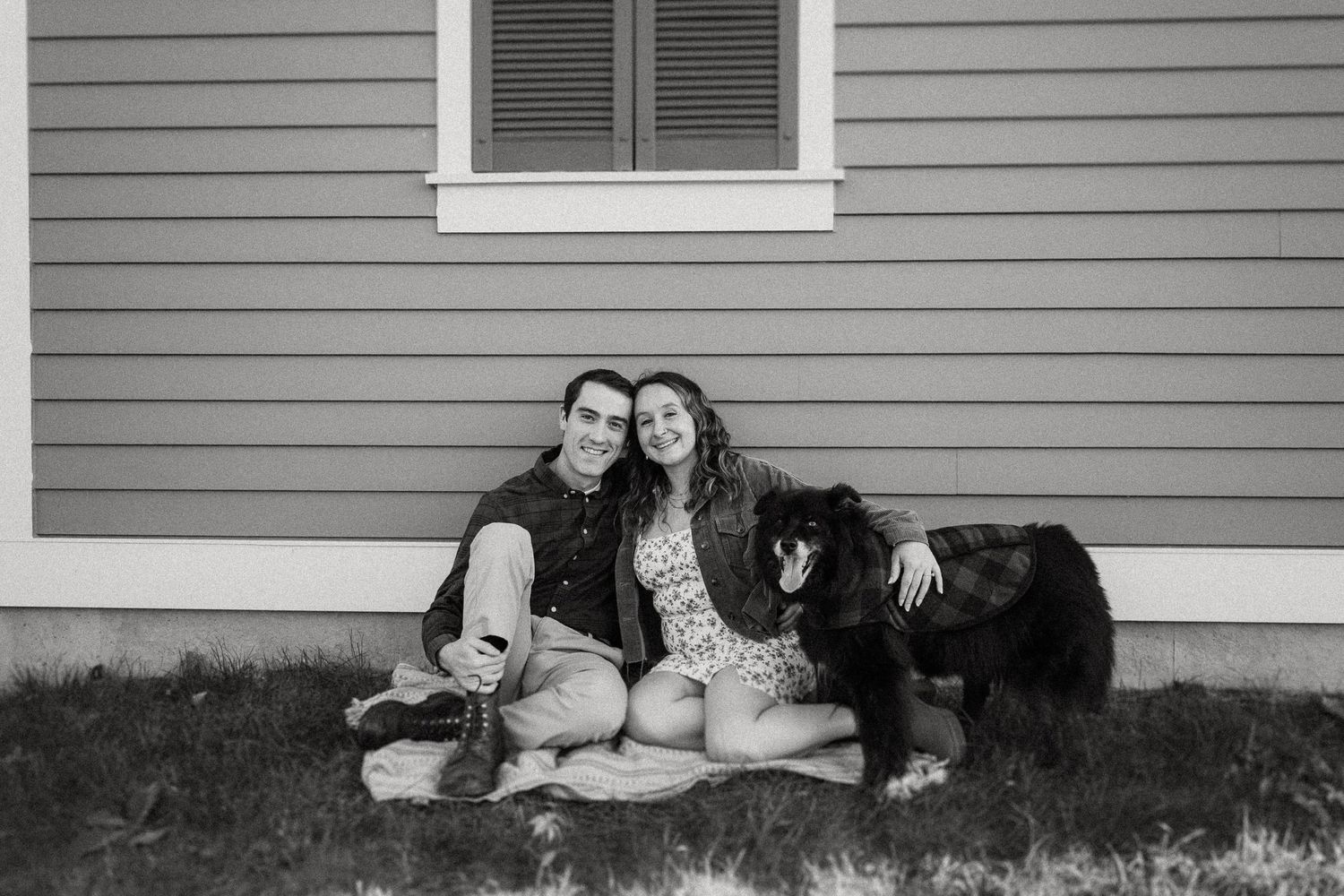 A black and white photo of a couple relaxing with their dog against a house exterior.