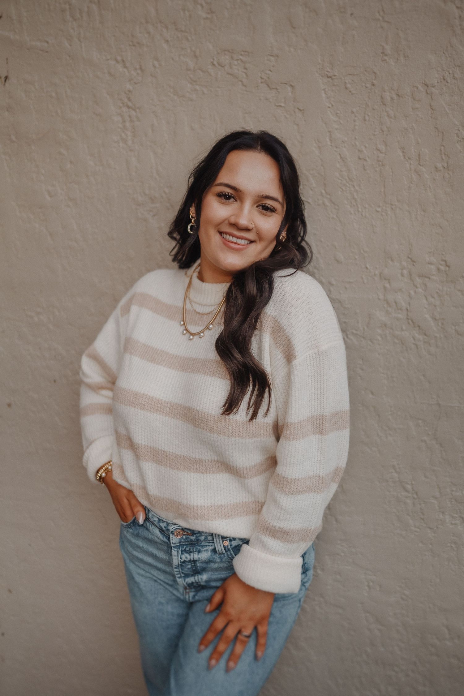 Casual portrait against beige wall featuring someone in striped sweater and jeans with layered necklaces.