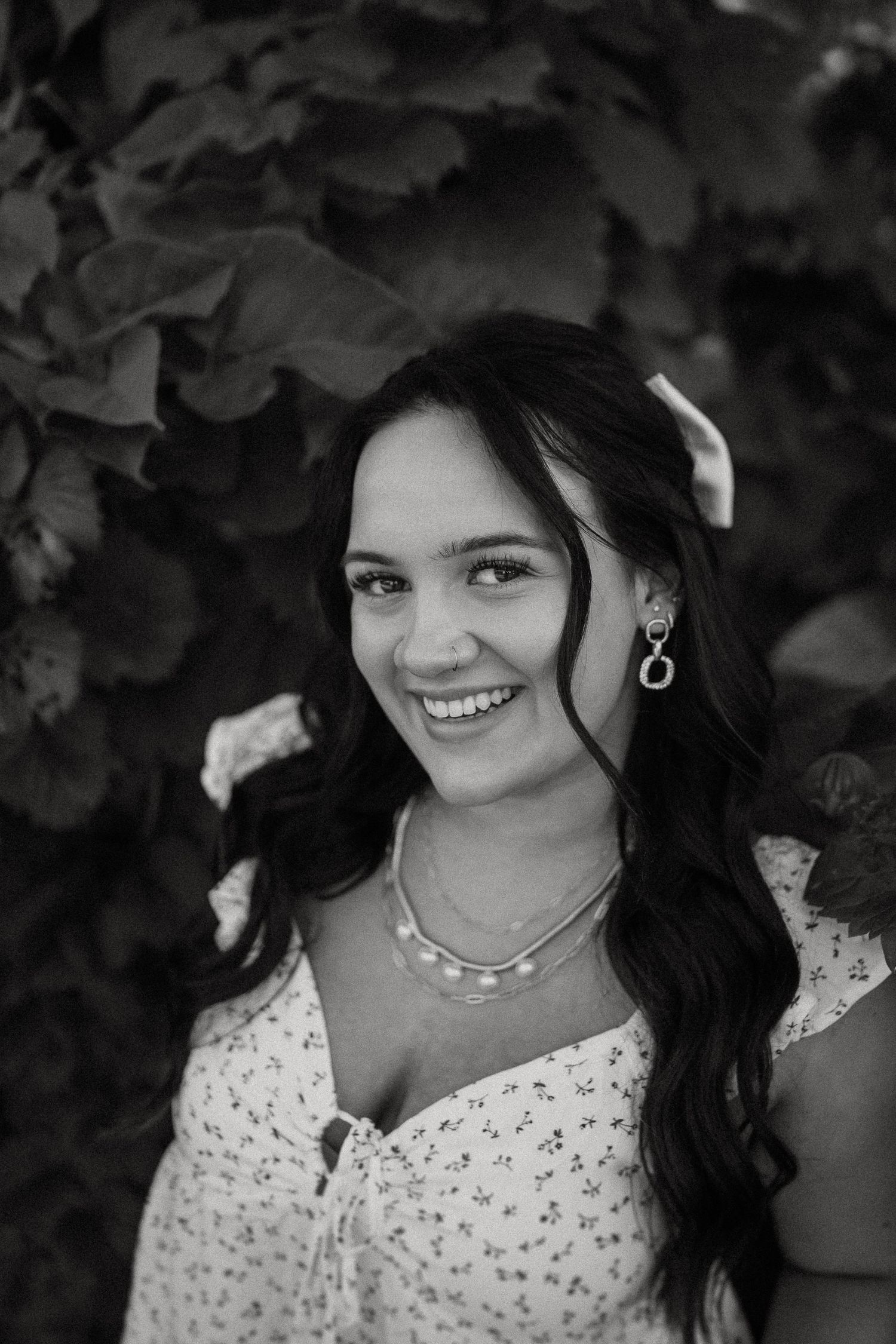 Black and white portrait of someone wearing a polka dot dress with pearl necklace and earrings against foliage background.