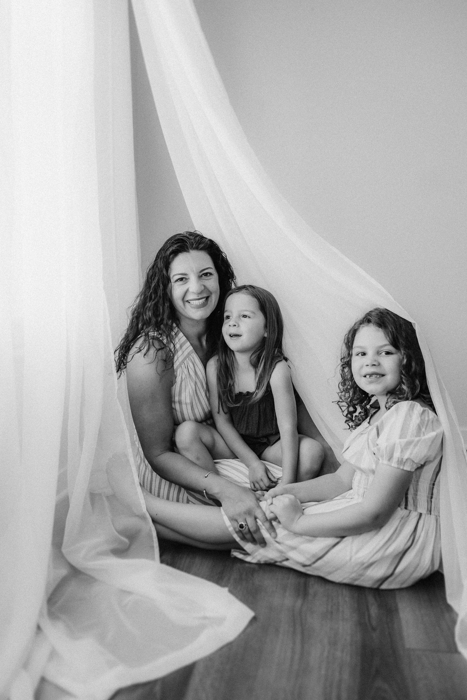 Black and white portrait under flowing white curtains on hardwood floor.