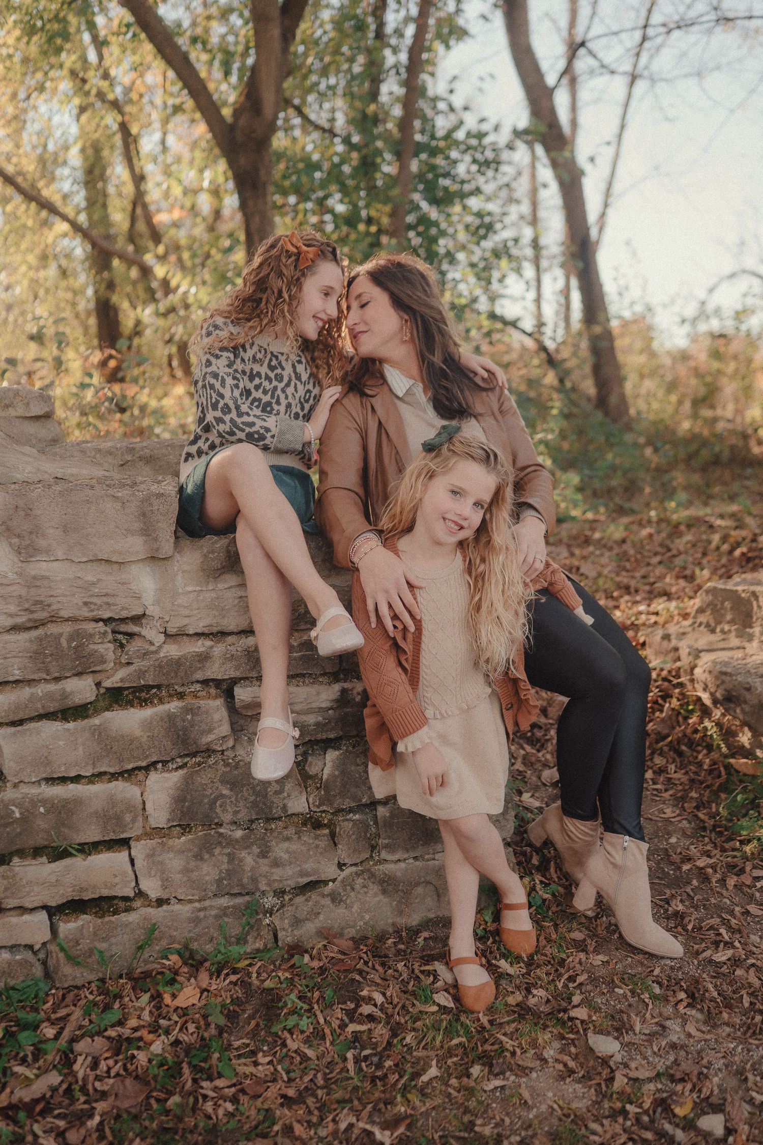 A couple shares intimate moments on a rustic stone wall surrounded by fallen autumn leaves in a wooded setting.