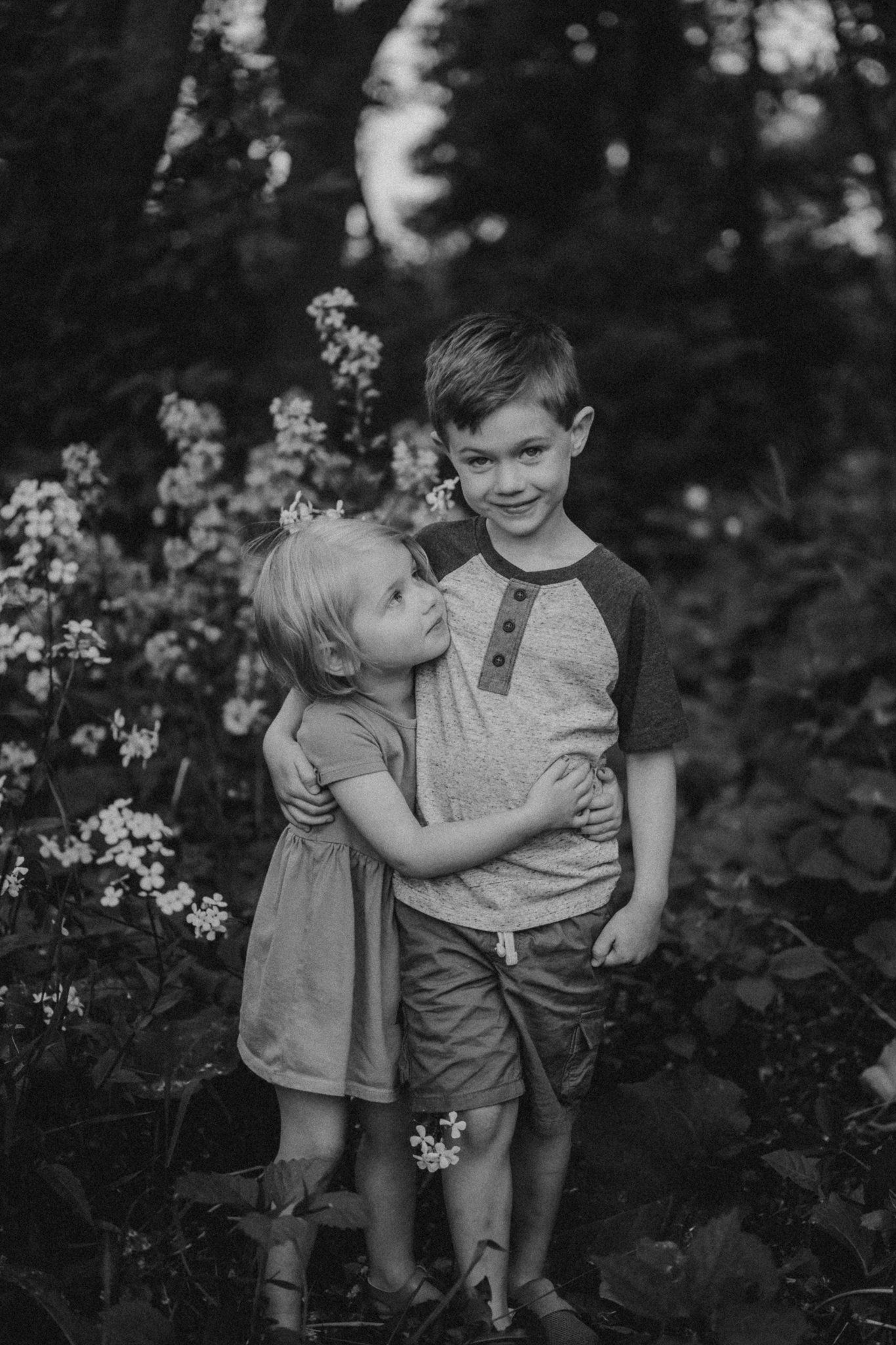 Black and white photo of siblings embracing among flowering plants in a garden.