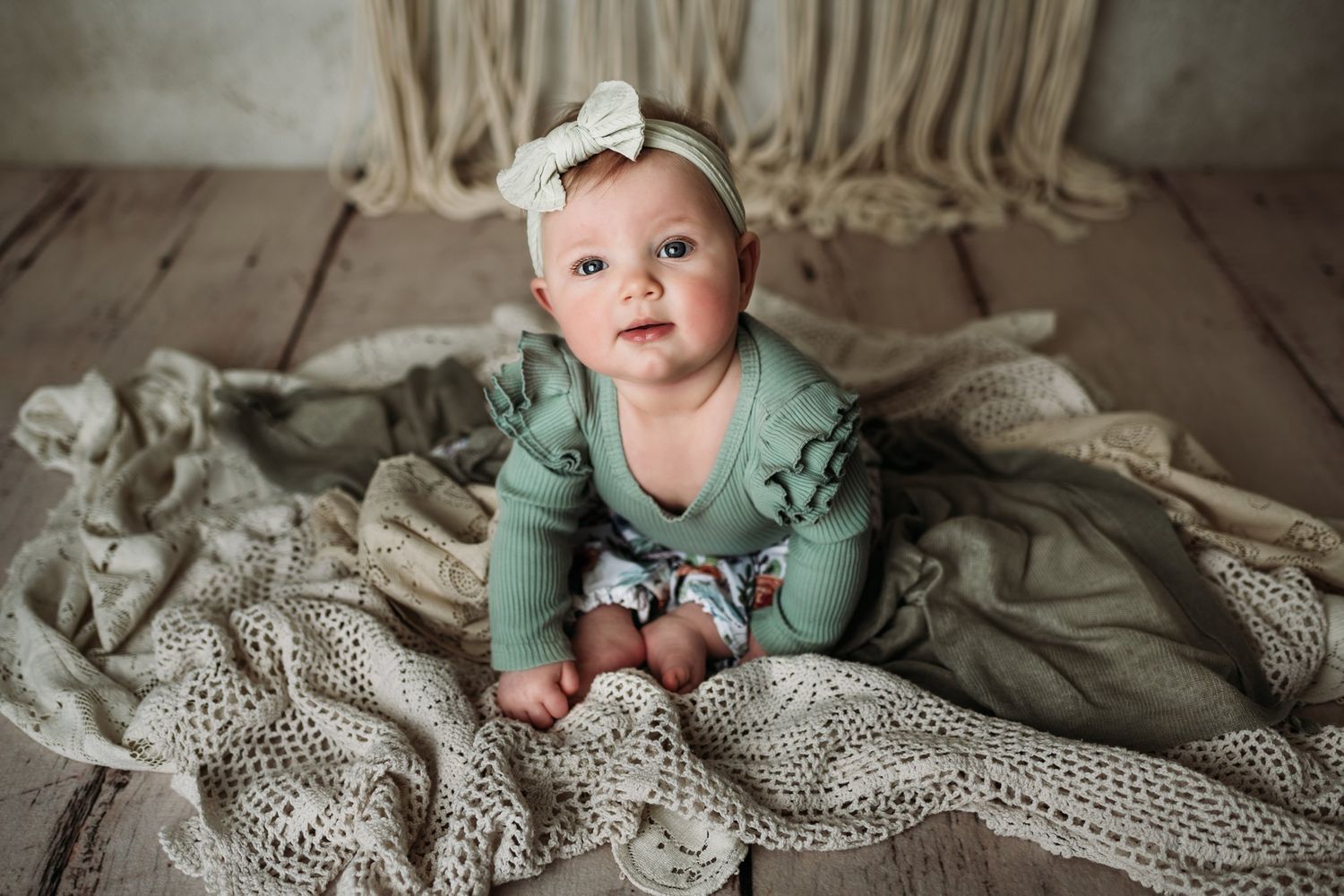 Baby in sage green outfit crawls on vintage lace blanket with macrame wall decor.