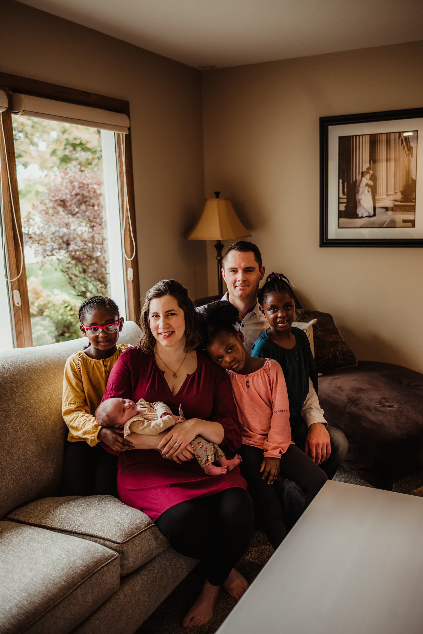 Family sitting together on living room couch with newborn baby near window.