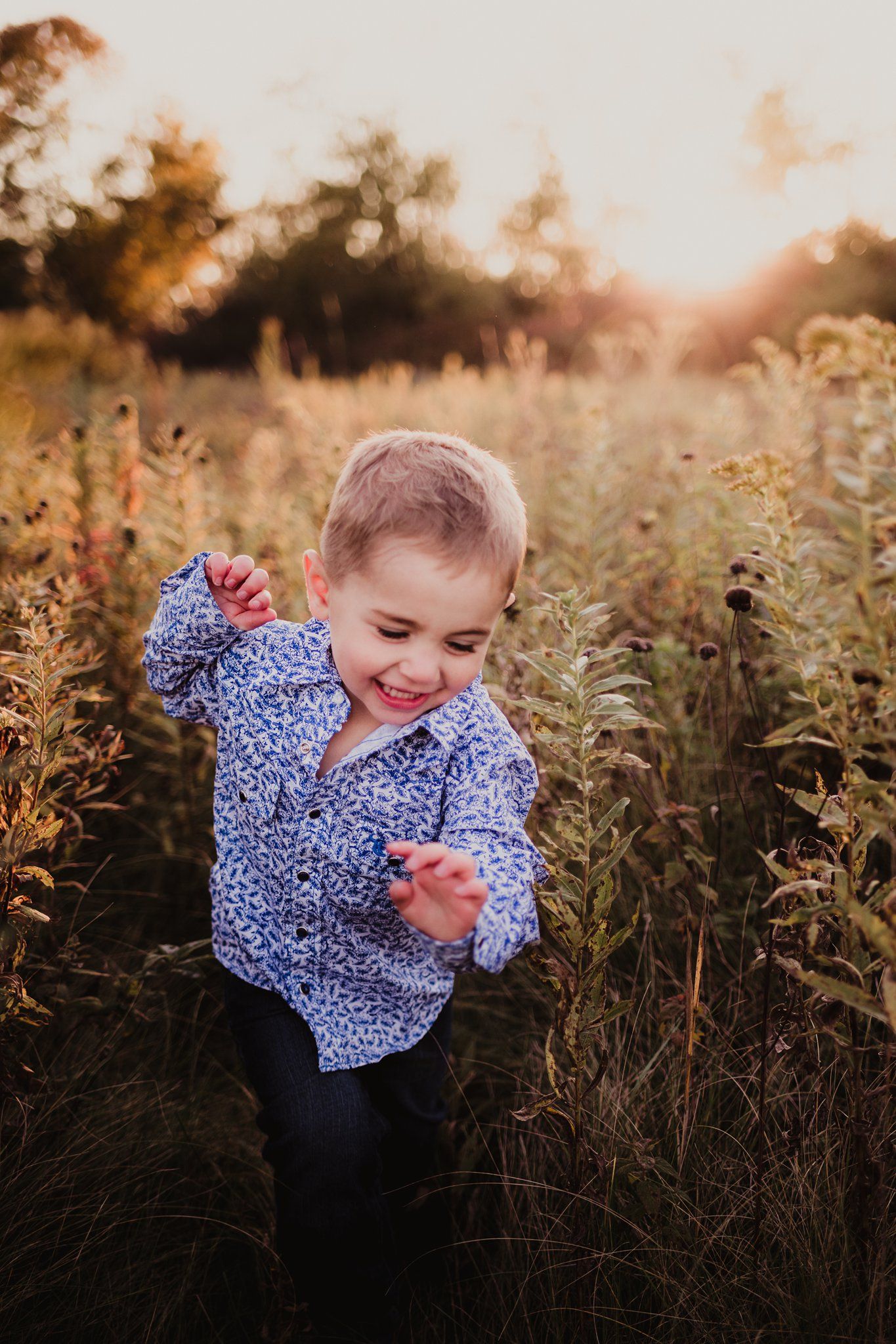 Young child in blue patterned shirt laughing and playing in field at golden hour.
