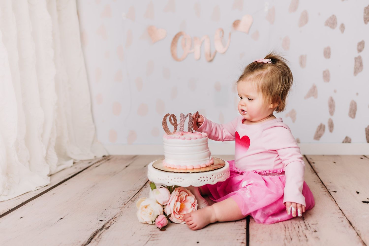 Pink-dressed figure sitting beside small birthday cake with floral decorations on wooden floor.