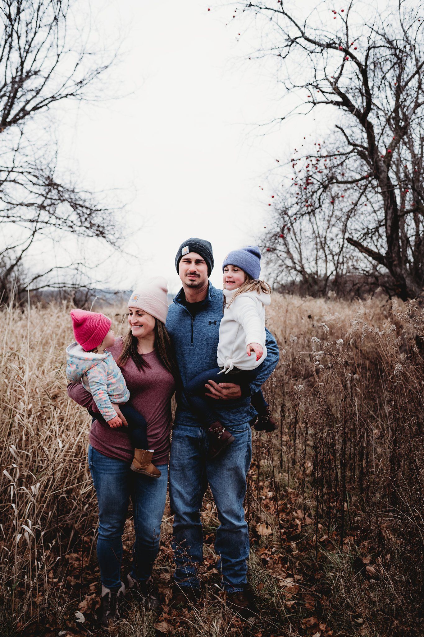 Family portrait in casual winter clothing standing among autumn grass and bare trees.