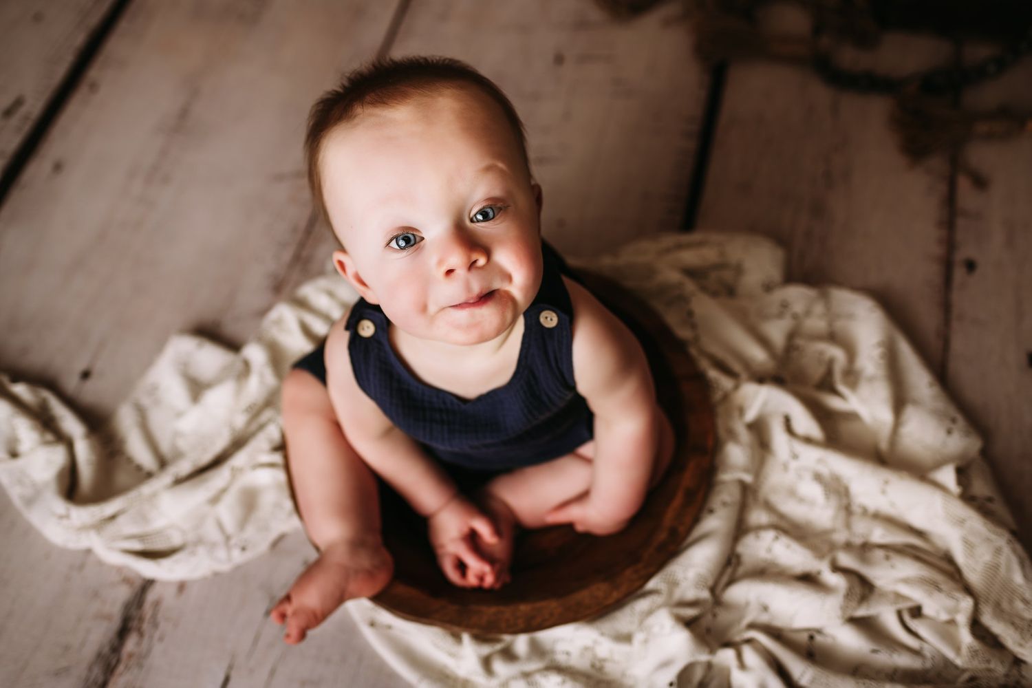 Baby in dark sleeveless outfit sits on wooden bowl against rustic floor and backdrop.