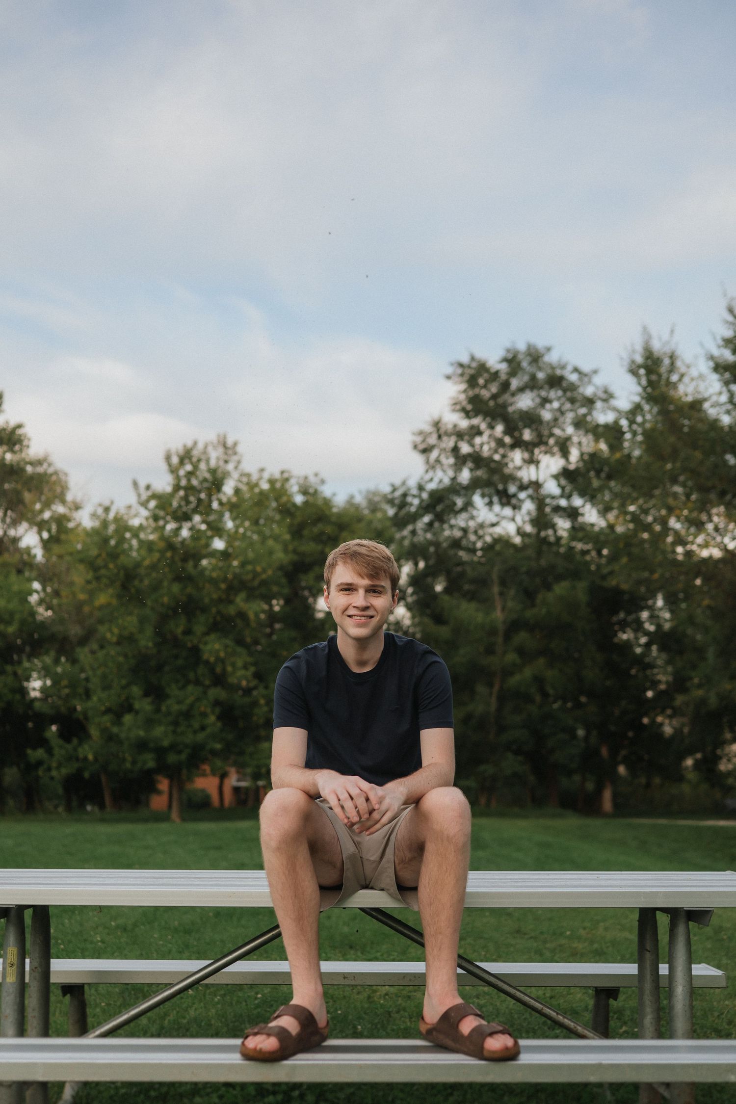 A person in a navy t-shirt sits on metal bleachers at a park or sports field on a sunny day.