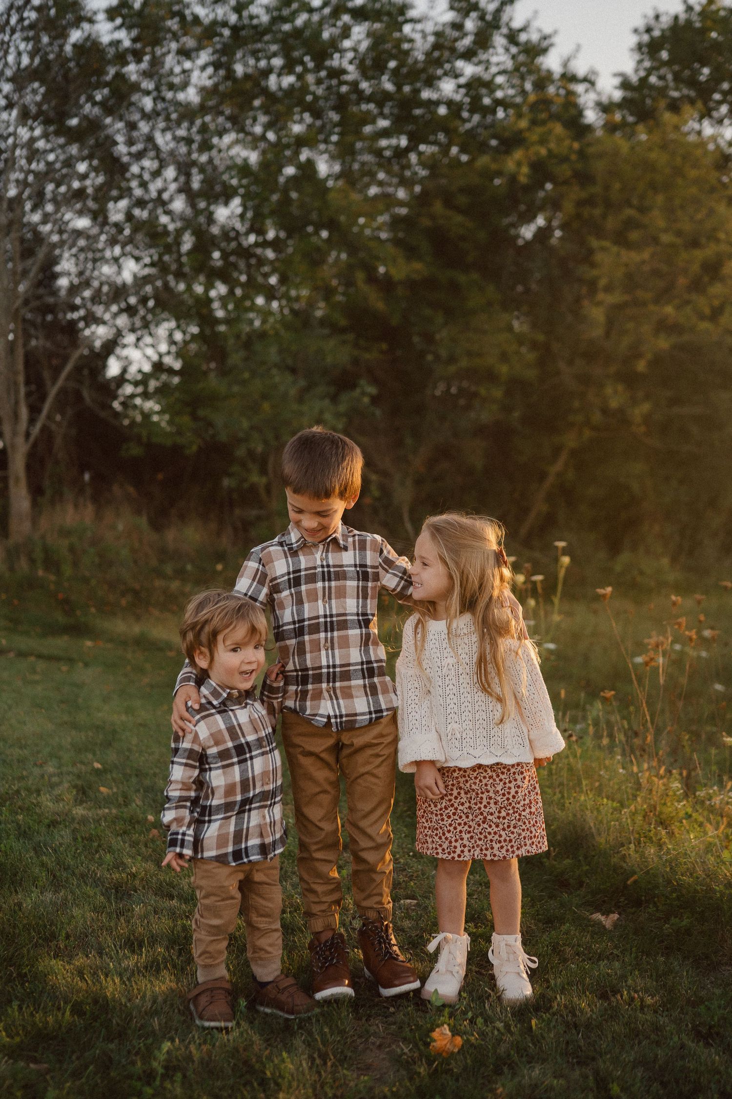 Three children sit together on a grassy hill at sunset wearing coordinated fall outfits in brown and plaid.