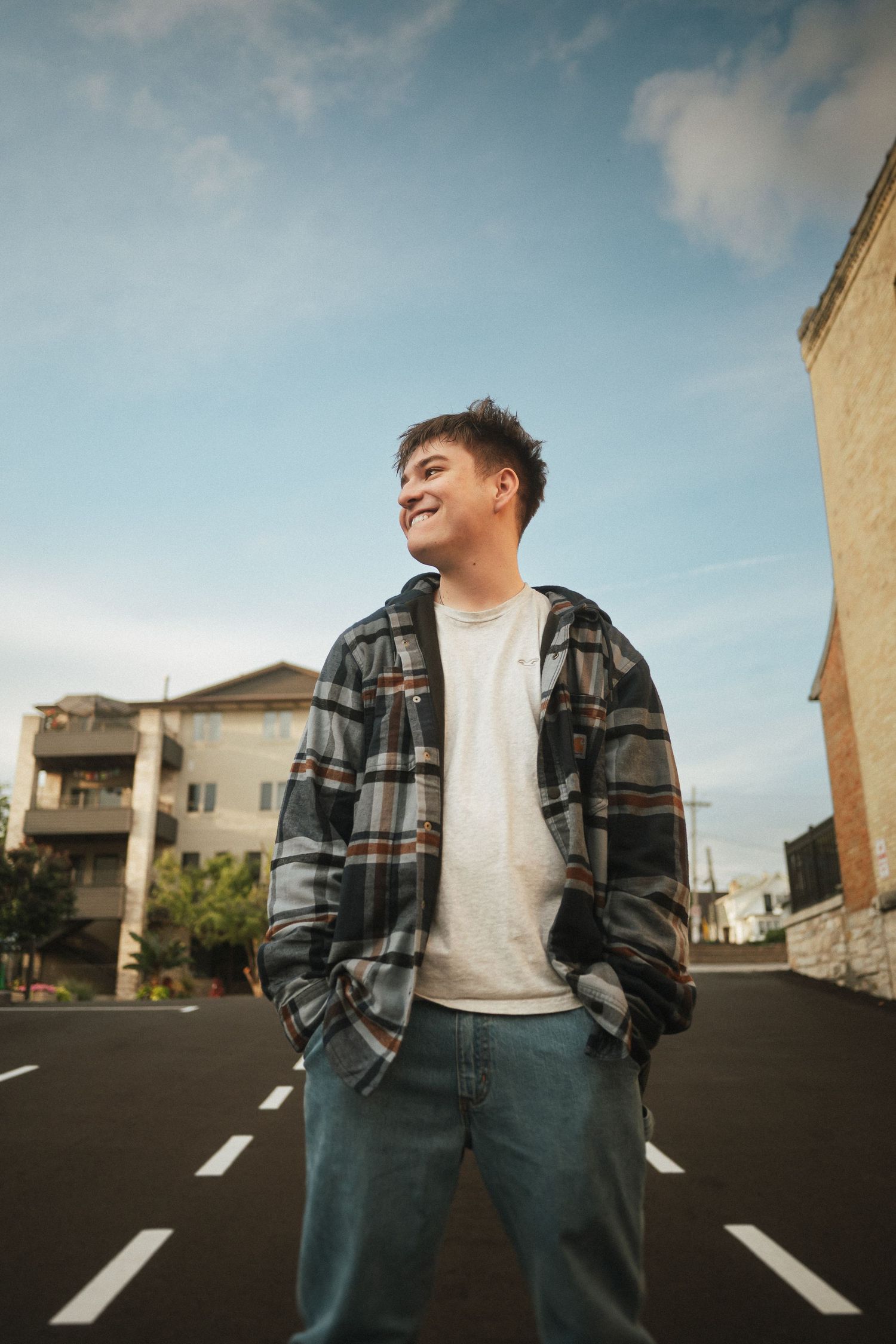 A person in a plaid jacket and white shirt poses in a parking lot against a cloudy sky.