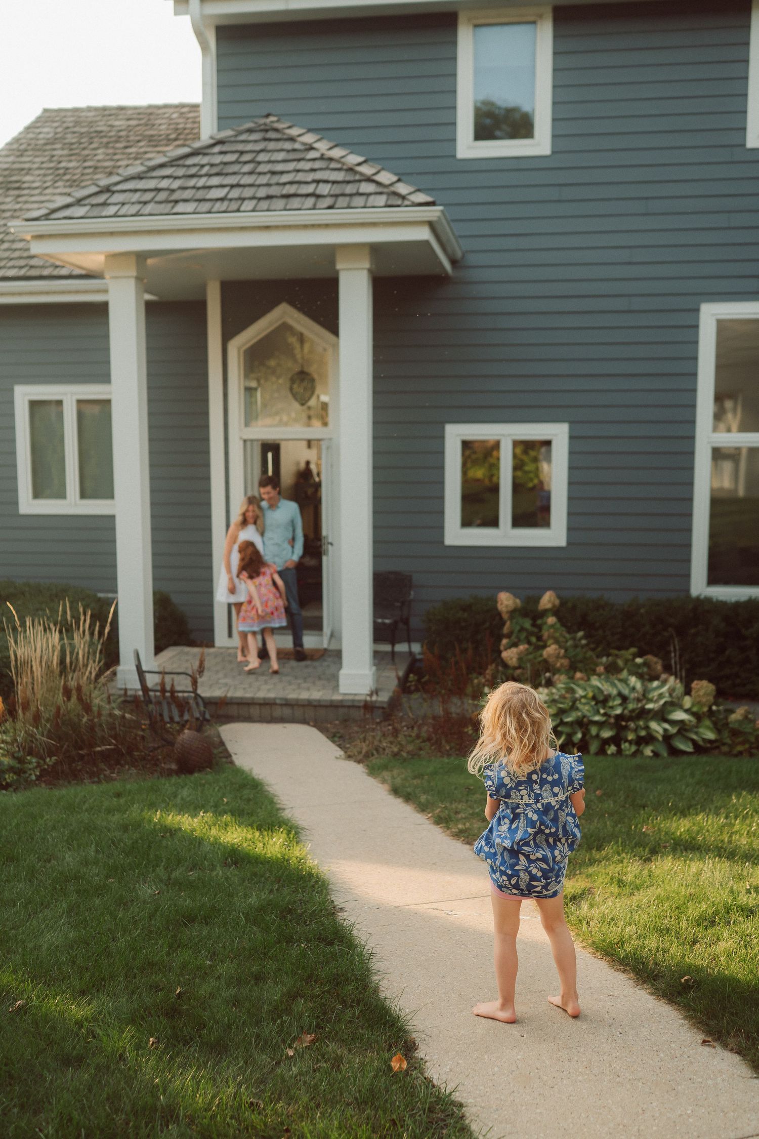 A sequence of walking along a concrete path leading to a blue-gray house with landscaped gardens.