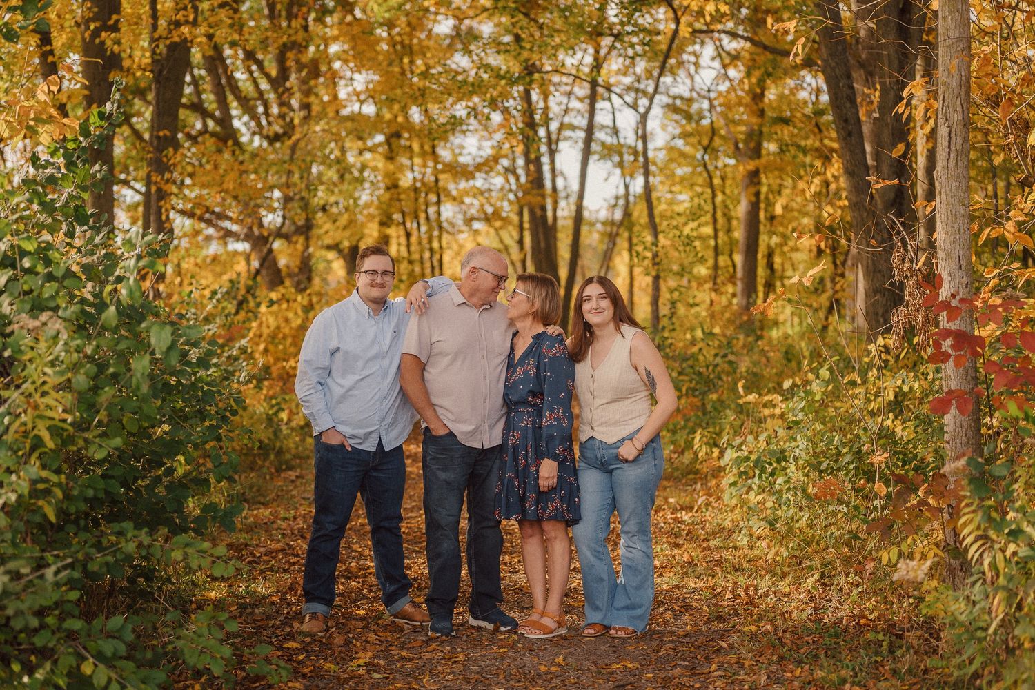 A group of three people pose together on a wooded path surrounded by golden autumn foliage.