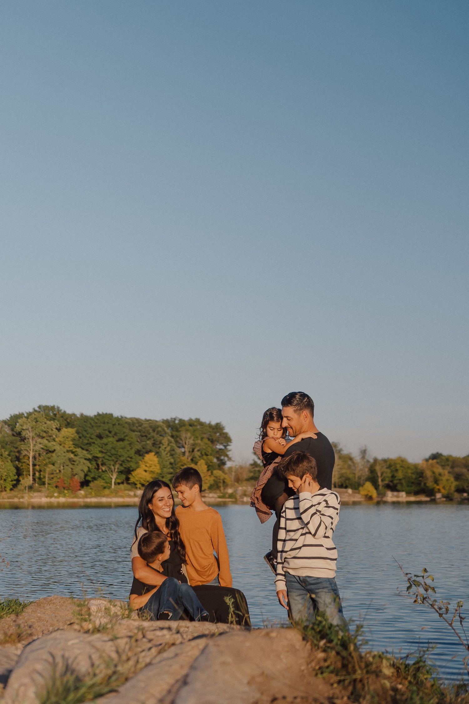 A couple shares an intimate moment by a scenic lake during sunset with trees in the background.