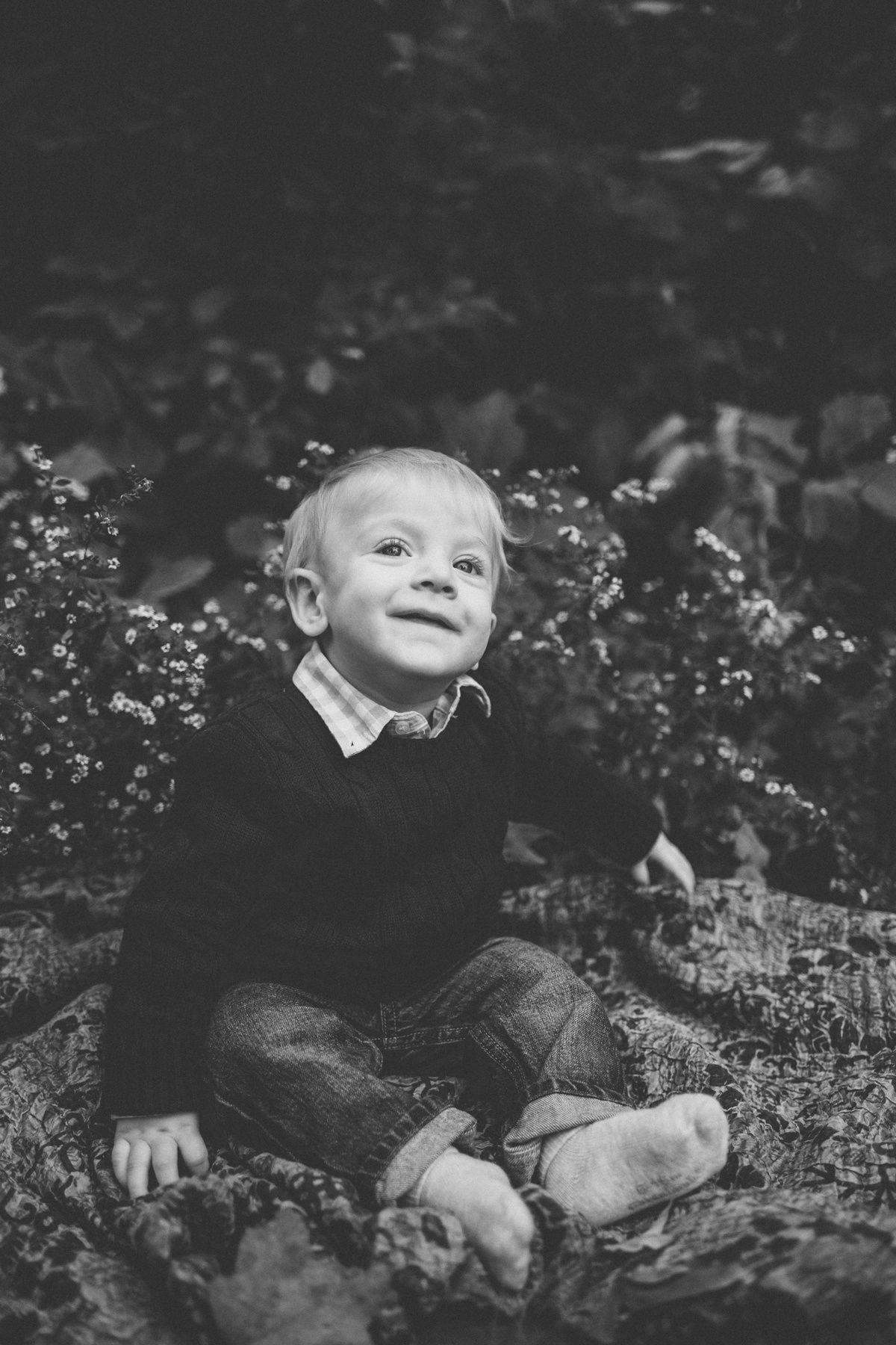 Black and white portrait of toddler sitting outdoors smiling among small flowers.