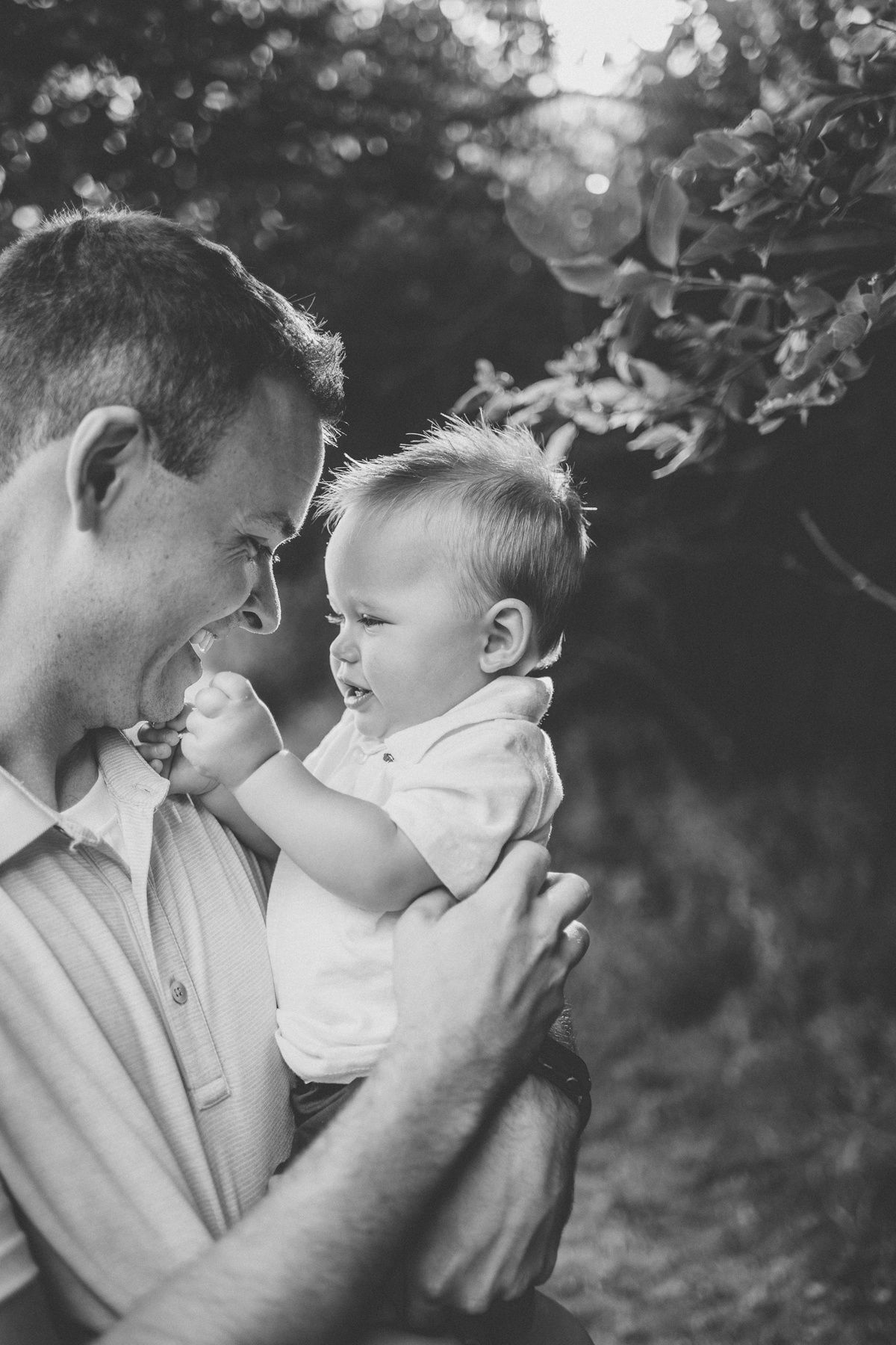 Black and white portrait of tender family moment outdoors with sun-lit bokeh background.
