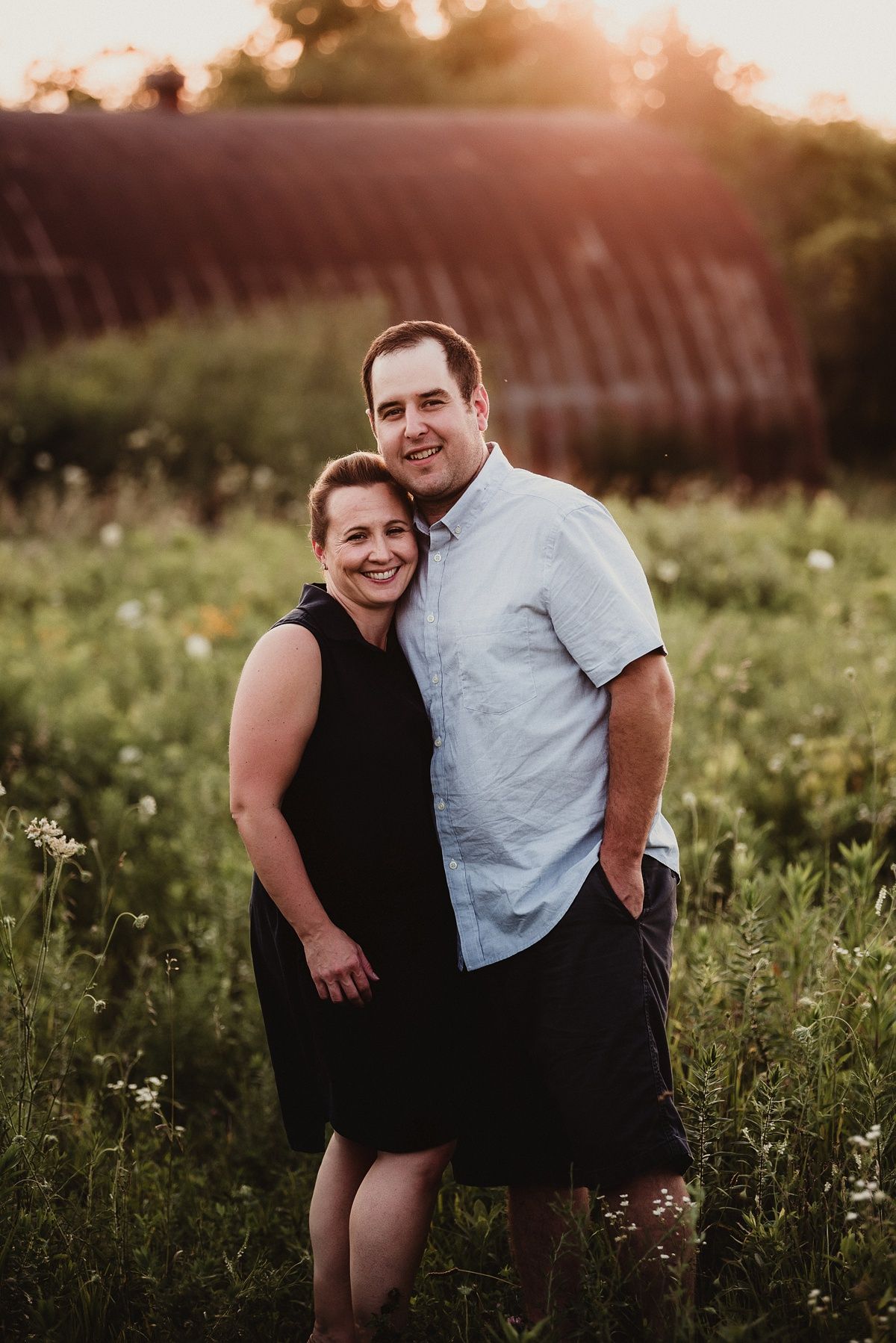 A couple poses together in a wildflower field at sunset with a rustic barn in the background.