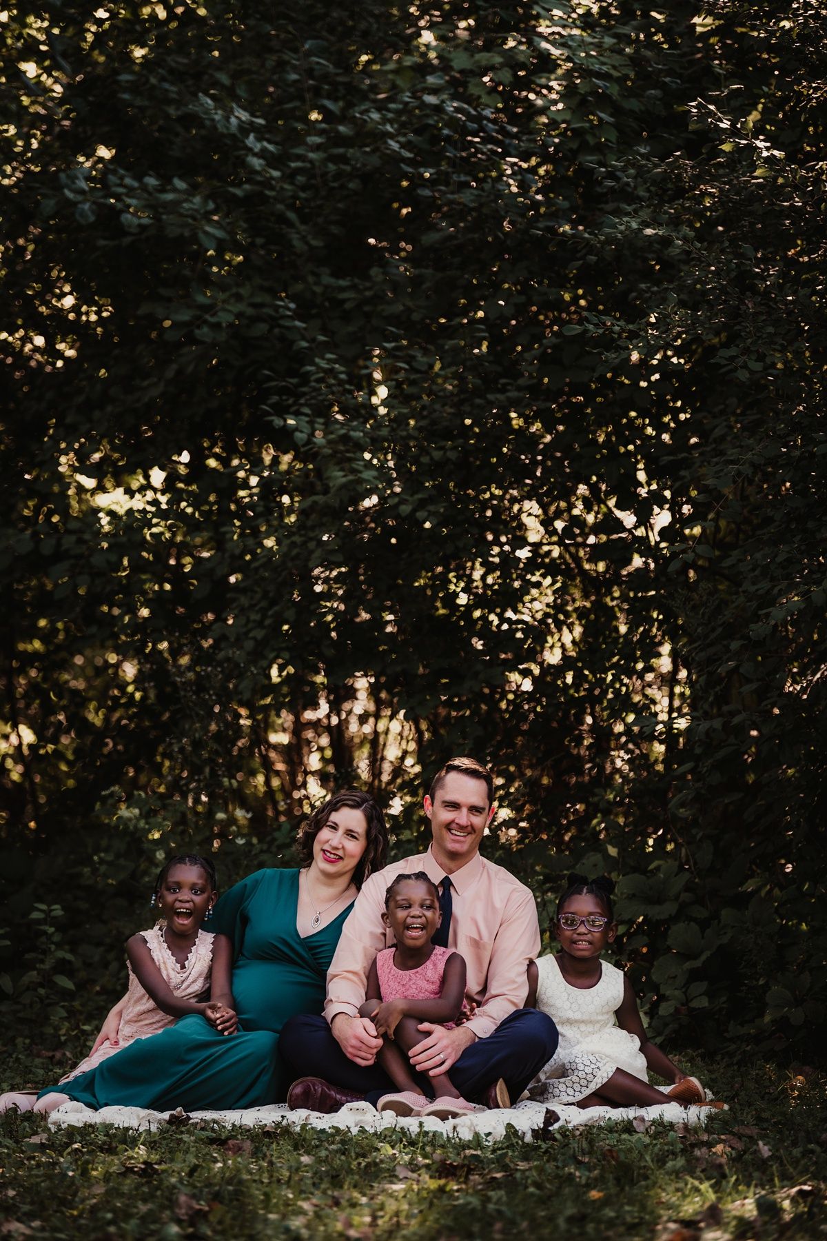 Family portrait on blanket in woods with golden sunlight filtering through trees.