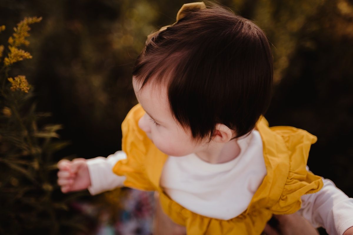 A toddler wearing a yellow pinafore dress looks to the side among autumn foliage.