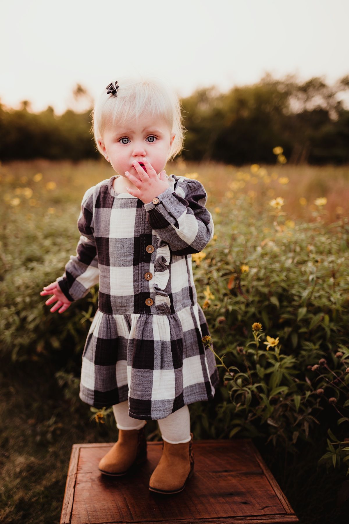 Small child in buffalo plaid dress and boots stands on wooden platform in field of wildflowers.