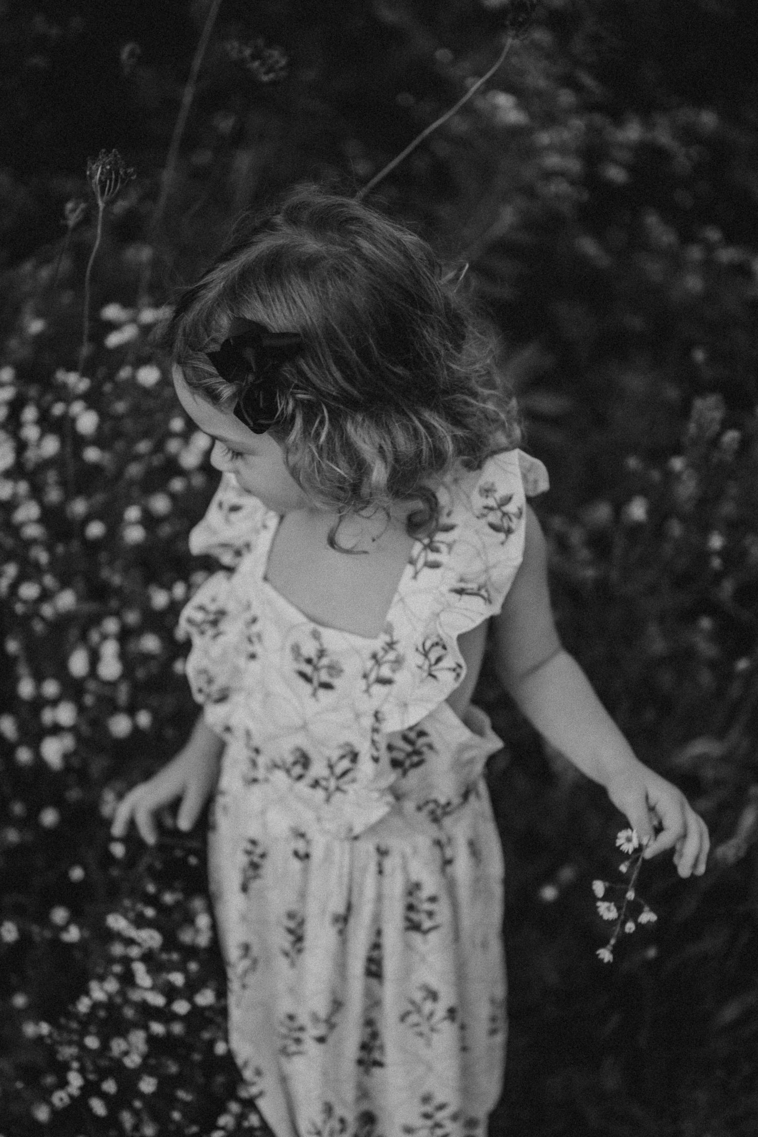 Black and white photo of child in floral dress dancing among wildflowers.