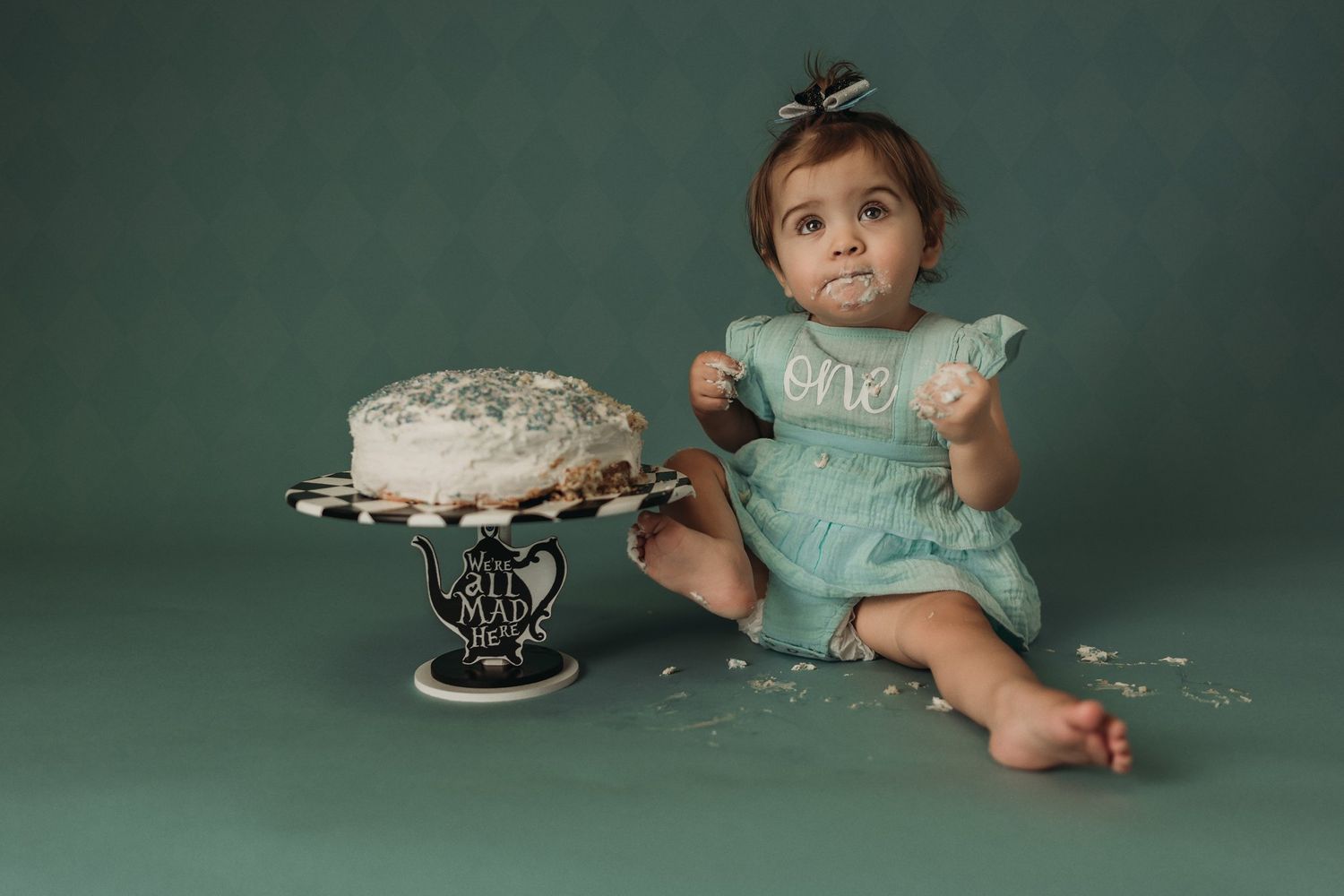 First birthday photo session with a white frosted cake on a decorative black stand against green backdrop.