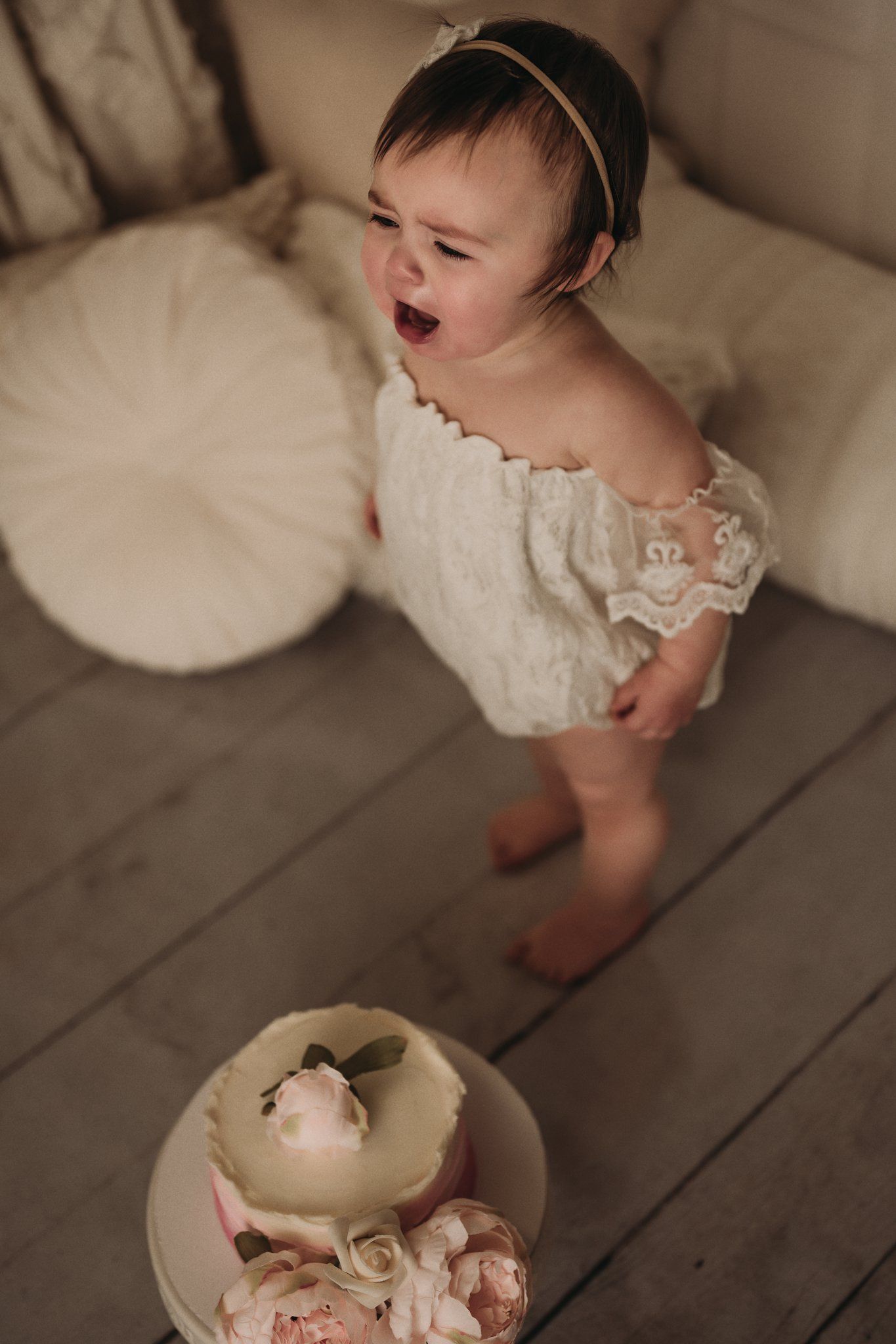 Baby in white lace outfit standing by pink cake with expressive yawning gesture.