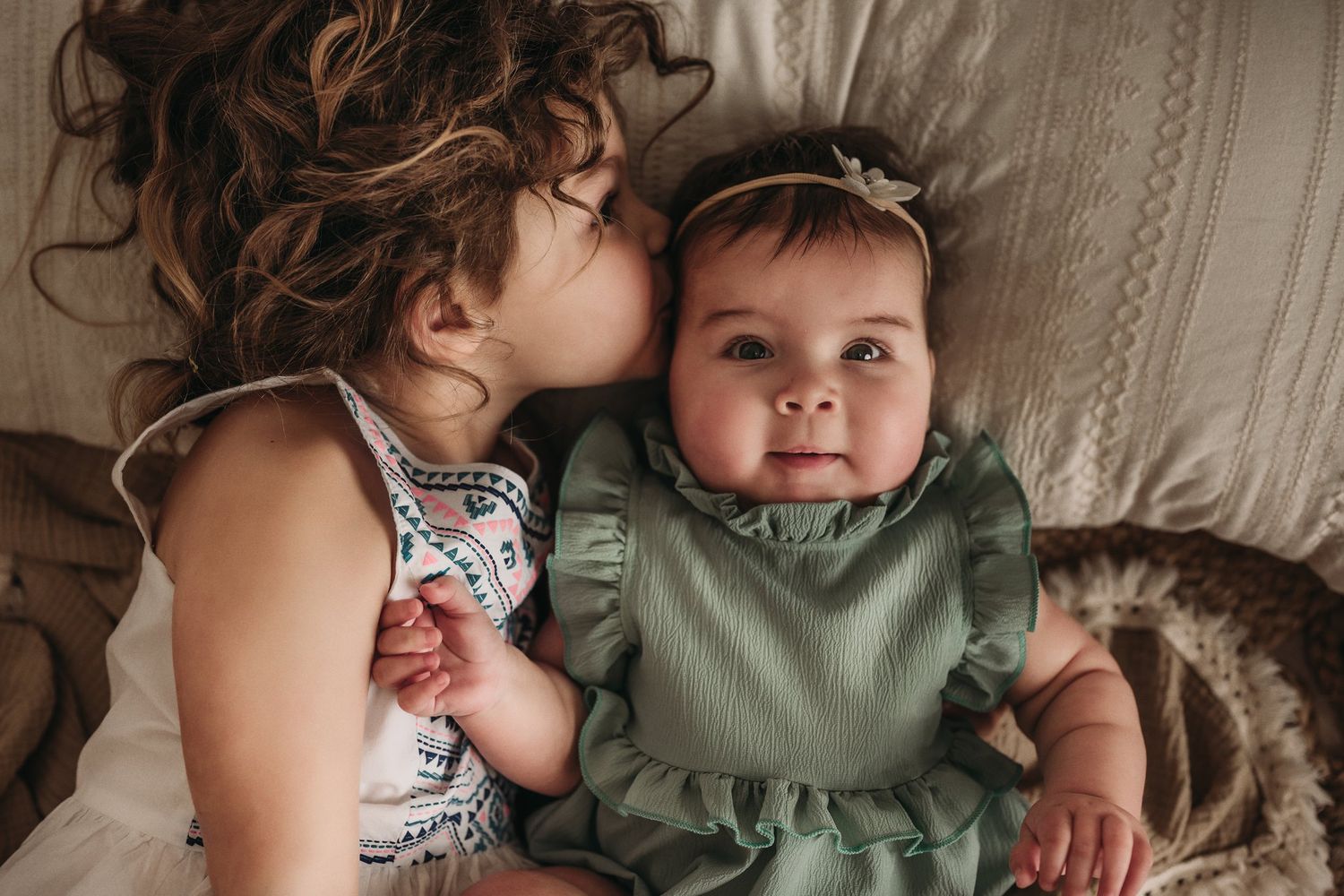 Two siblings share a tender moment while laying on a cream-colored couch with textured cushions.