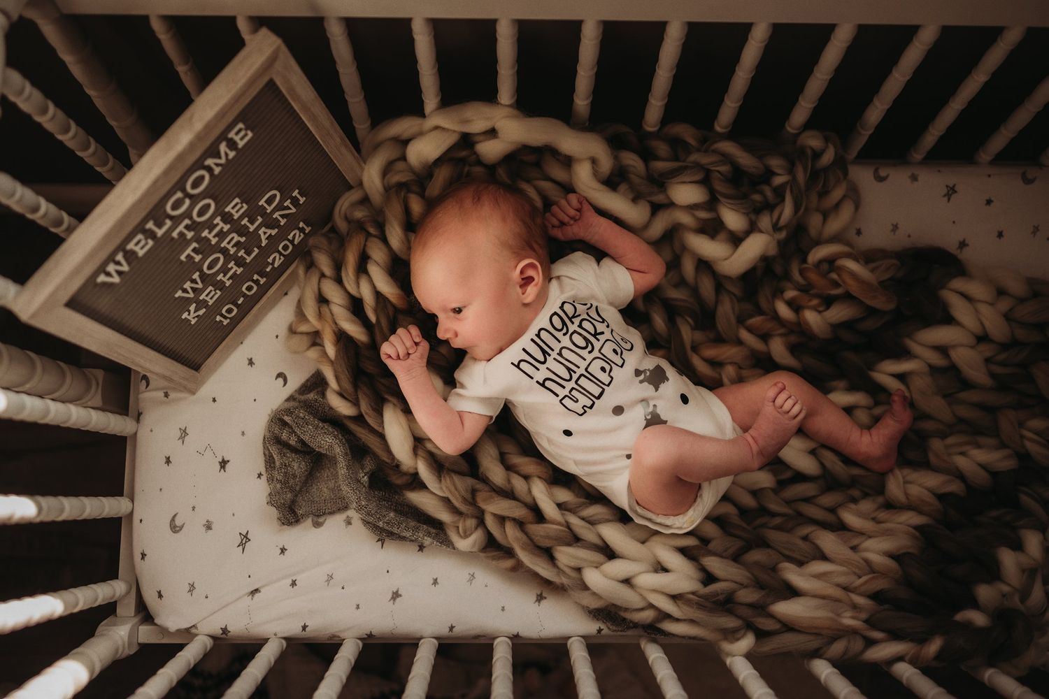 A sleeping infant rests in a wooden crib with chunky knit blanket and milestone cards.