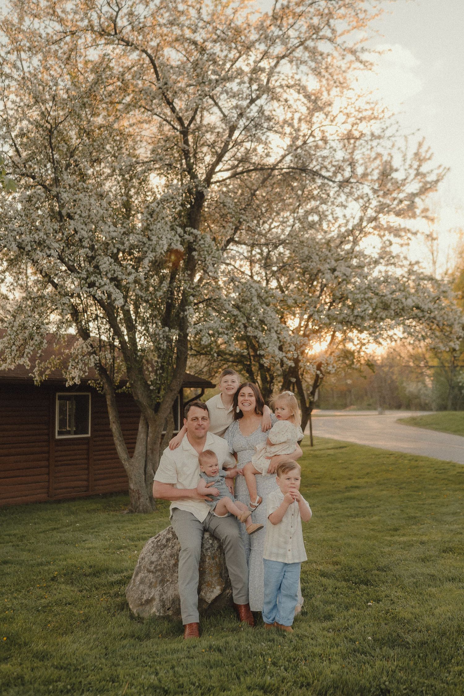 Happy family of six posing together under a blooming white tree at golden hour in a spring garden.