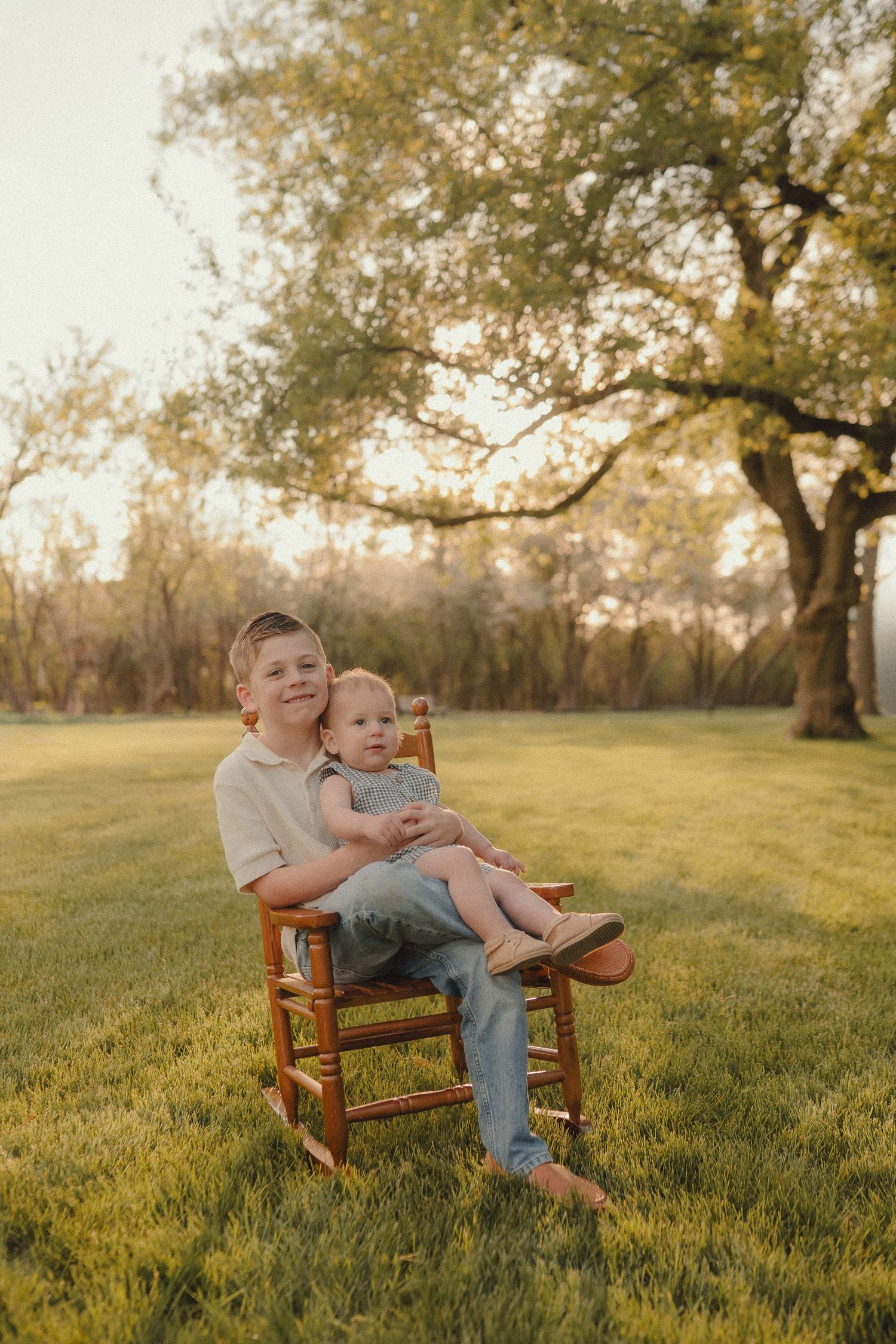 Boy sits in wooden chair holding baby sibling in sunny green park with large tree backdrop.