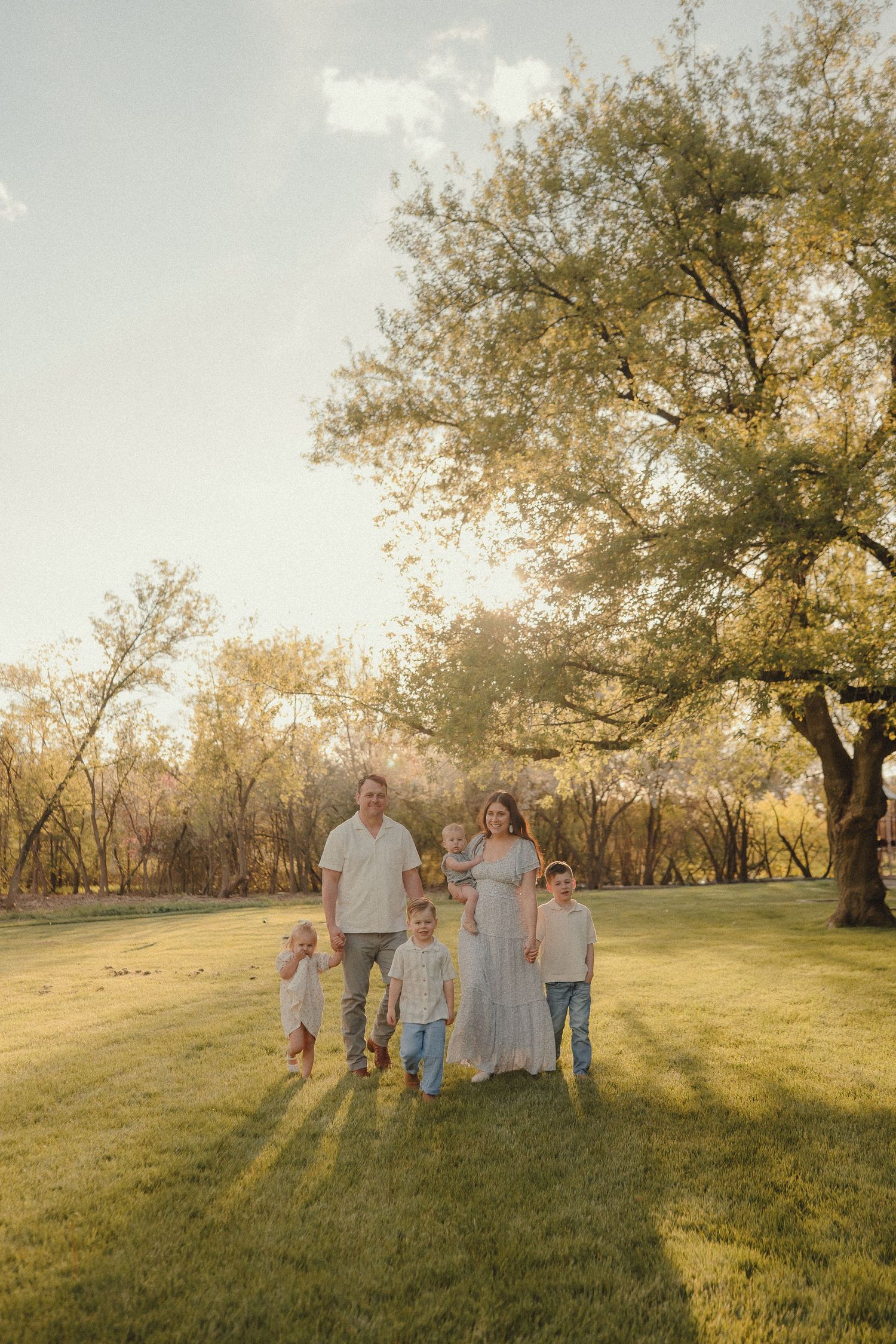 Family of six walking through a sunny green park with large trees in golden hour light.