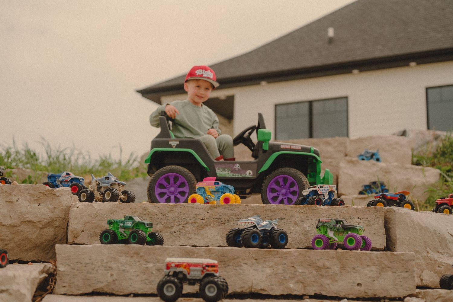 Boy in red cap sits on green ride-on jeep surrounded by monster truck toys on stone steps.