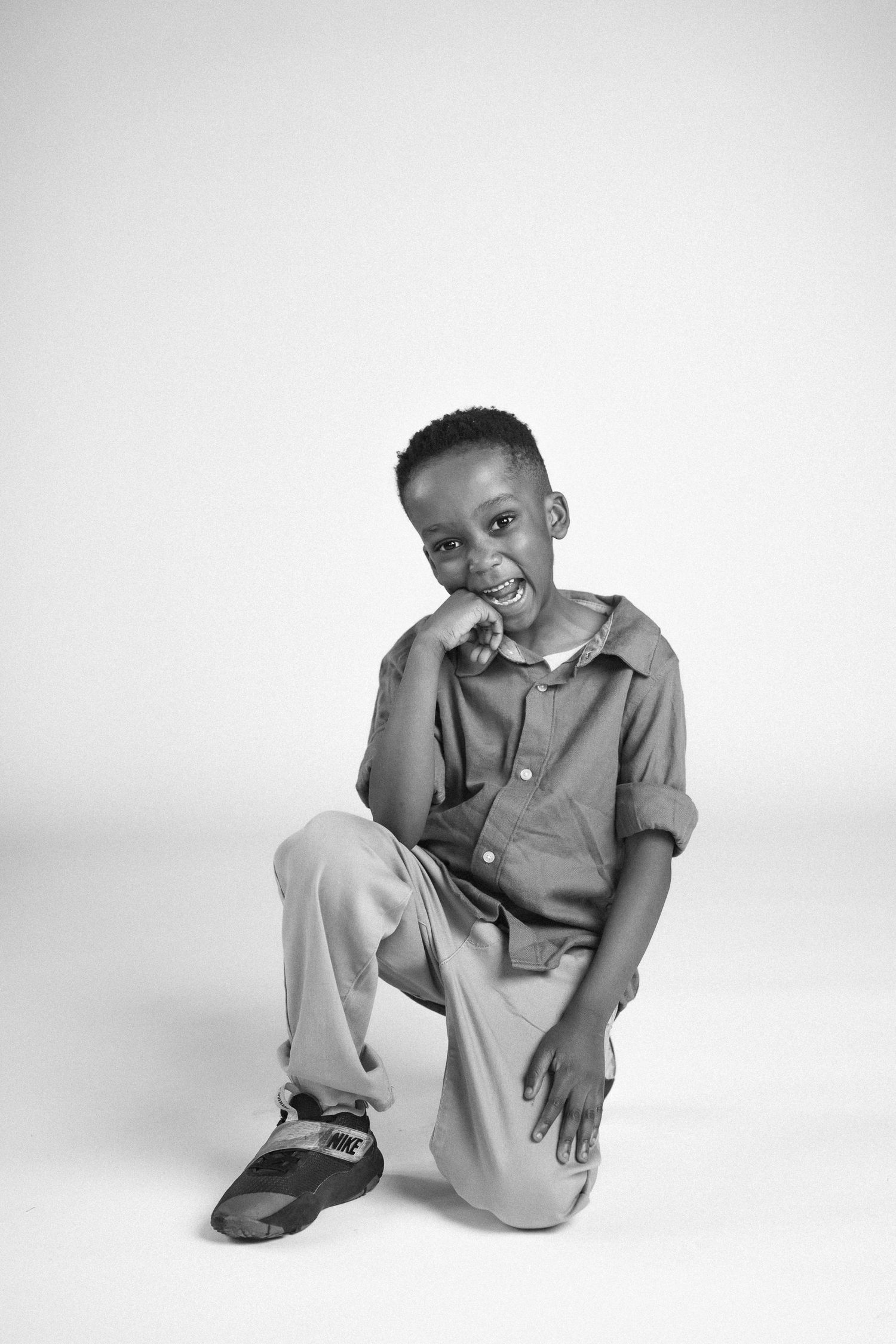 A young child in casual clothing kneels while smiling against a white studio background.