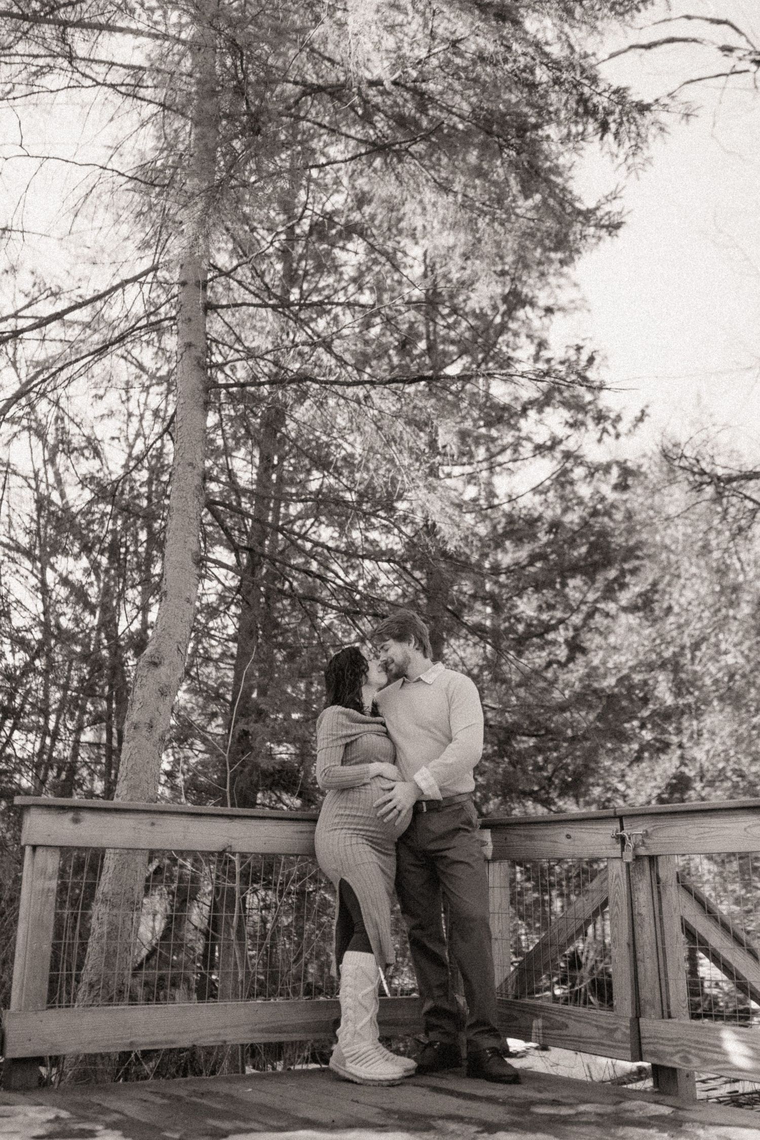 A couple shares an intimate moment on a wooden deck surrounded by tall pine trees in black and white.