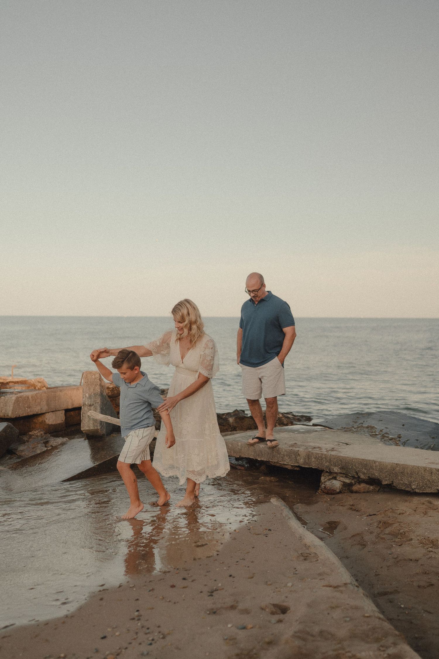 A family enjoys a playful moment on a sandy beach at sunset, with a woman in white, a boy, and a man.