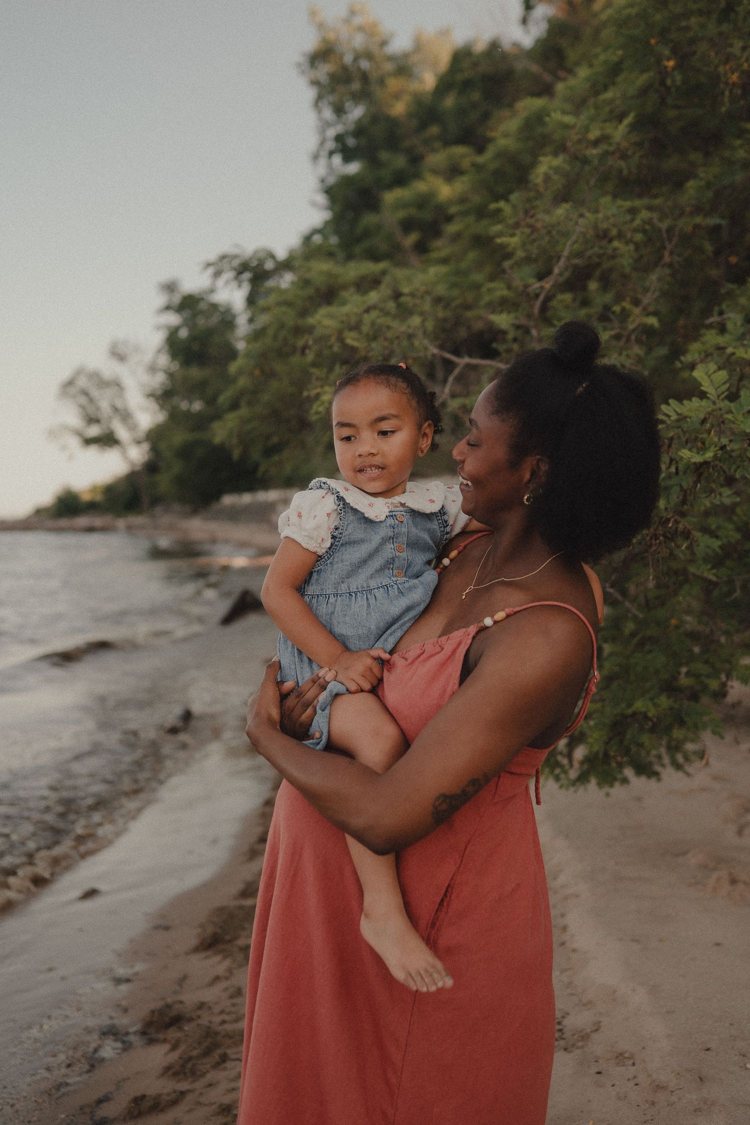 Pregnant Black mother in coral dress tenderly holding young toddler daughter on sandy beach at dusk.
