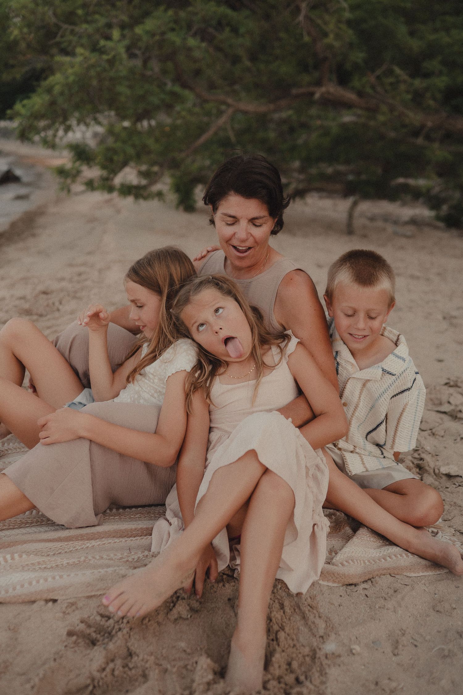 Laughing mother sits on beach blanket with three playful children making silly faces outdoors.