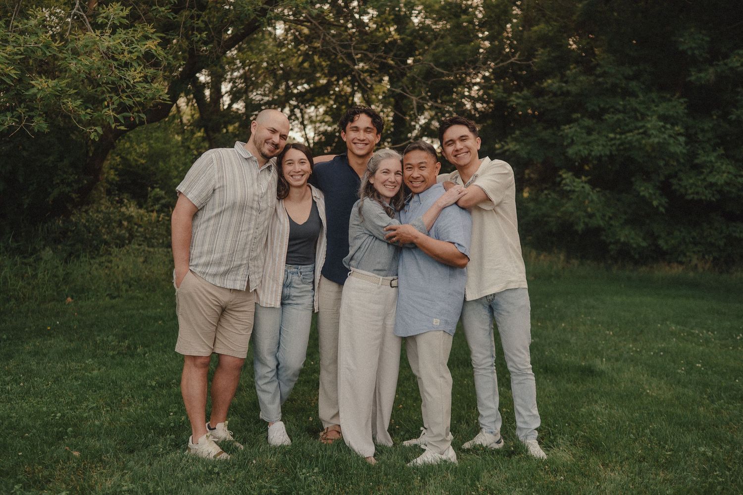 A joyful family of six poses together outdoors on green grass with lush trees in the background.