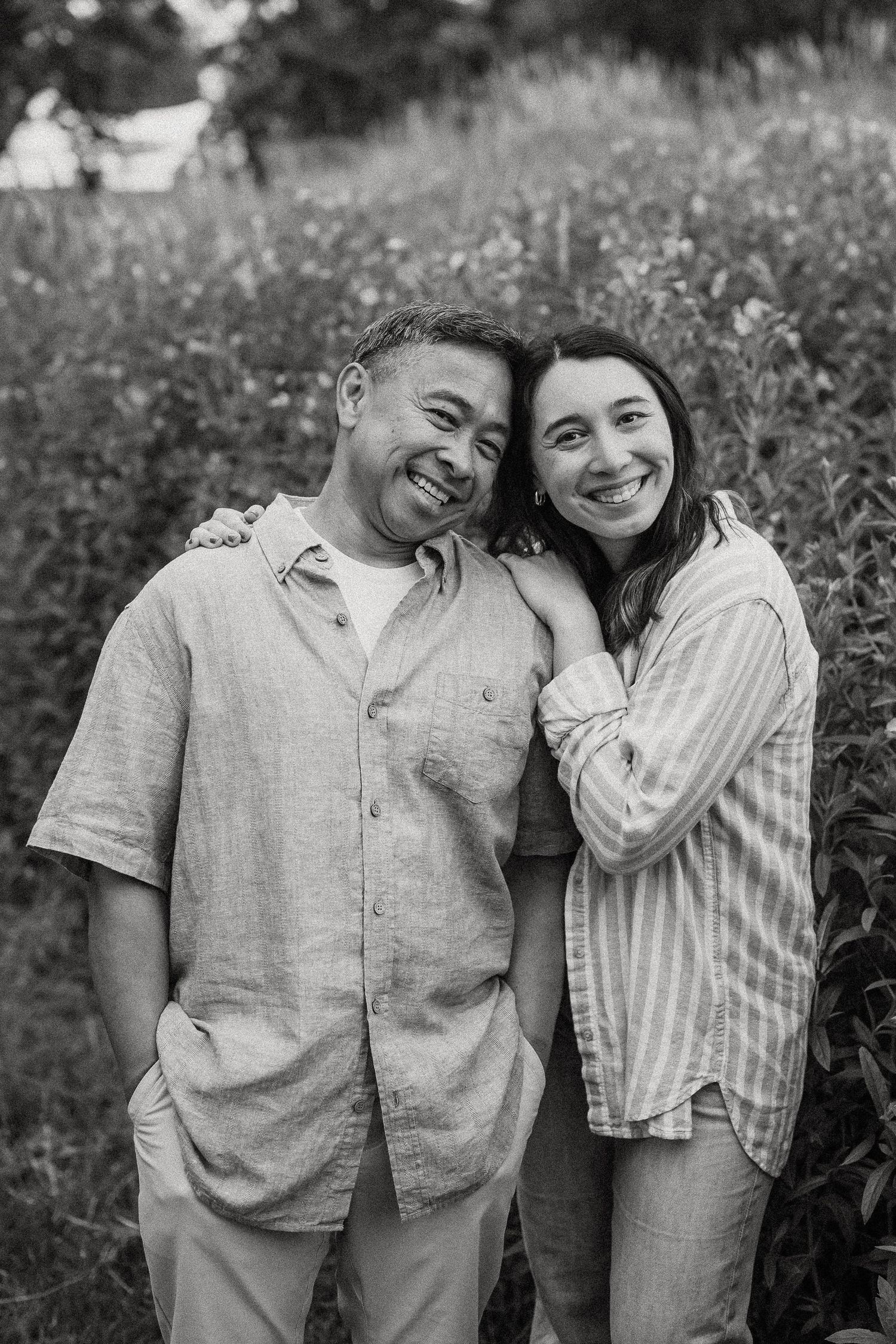 Black and white photo of a smiling couple embracing outdoors in a lush green field setting.