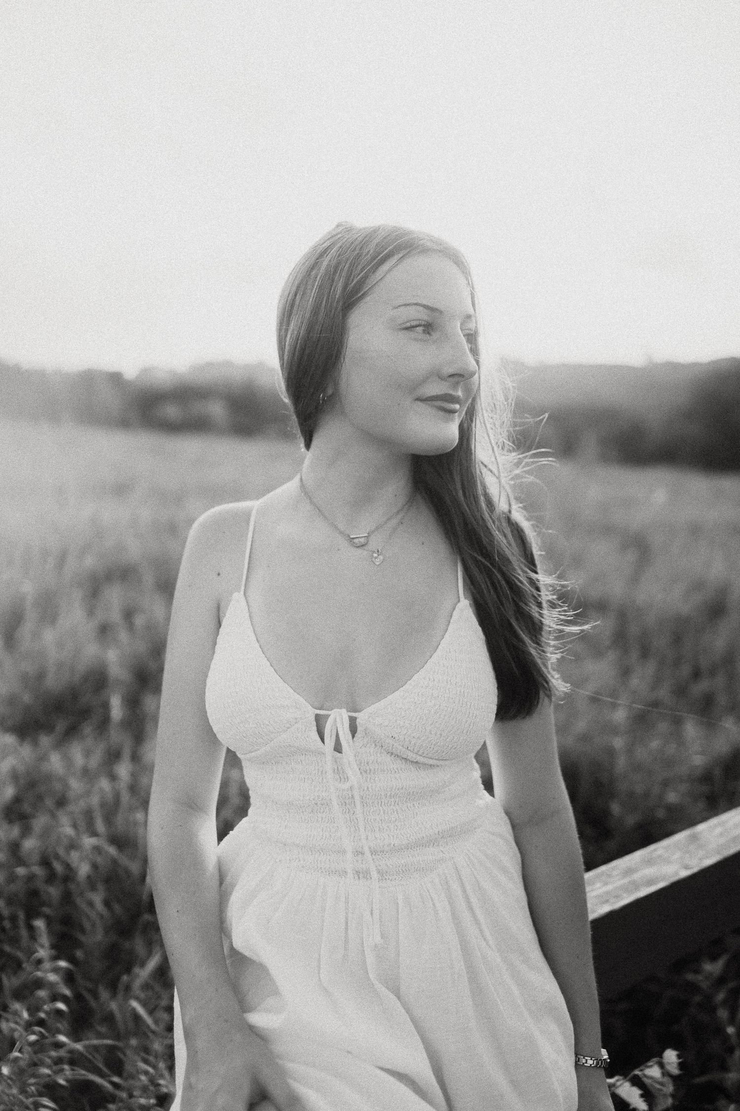 Black and white portrait of young woman in white sundress standing in a field at sunset.