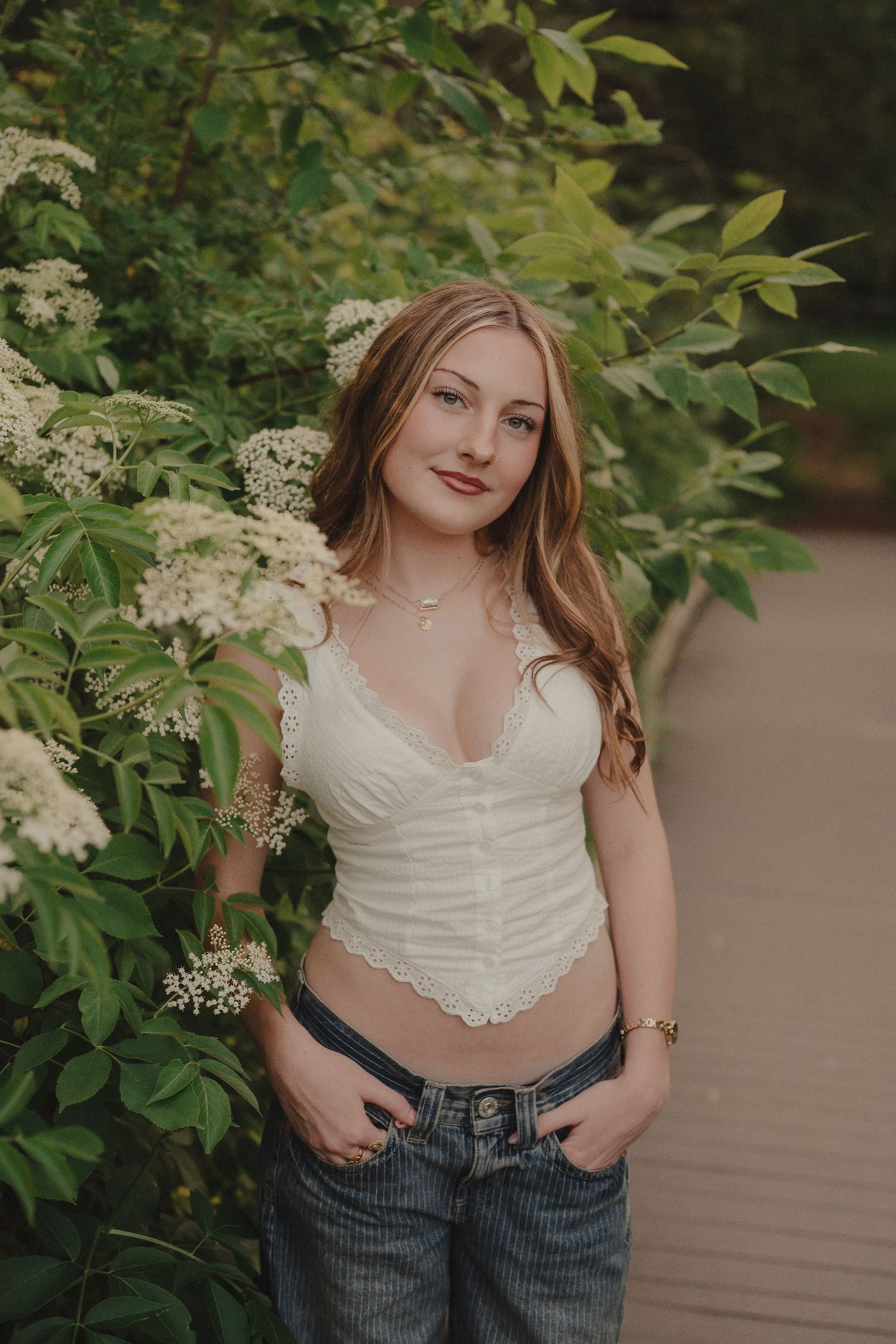 Young woman in white lace crop top and jeans posing beside blooming white flowers in a lush green garden.