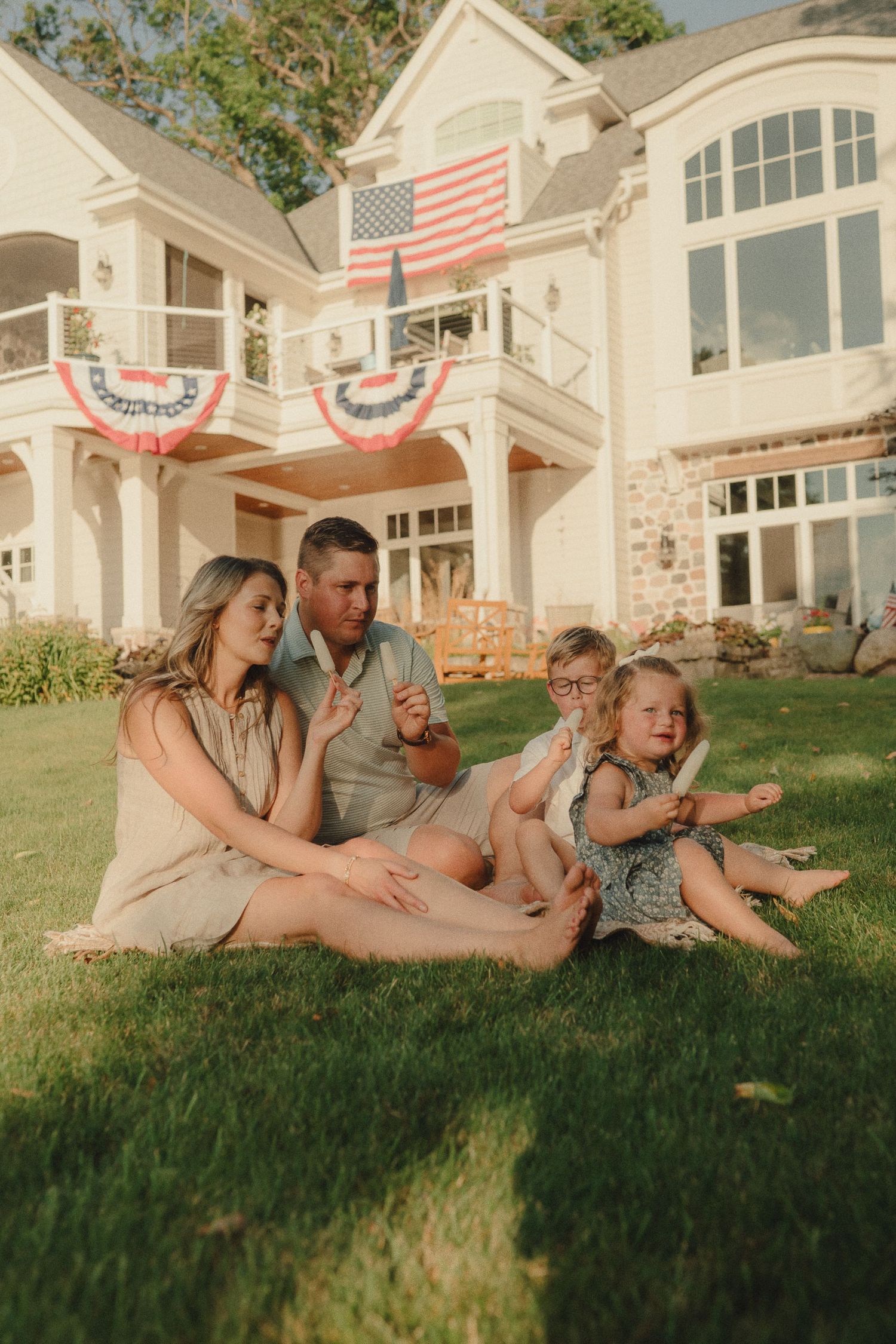 Family of four sitting on lawn in front of large home decorated with American flags.